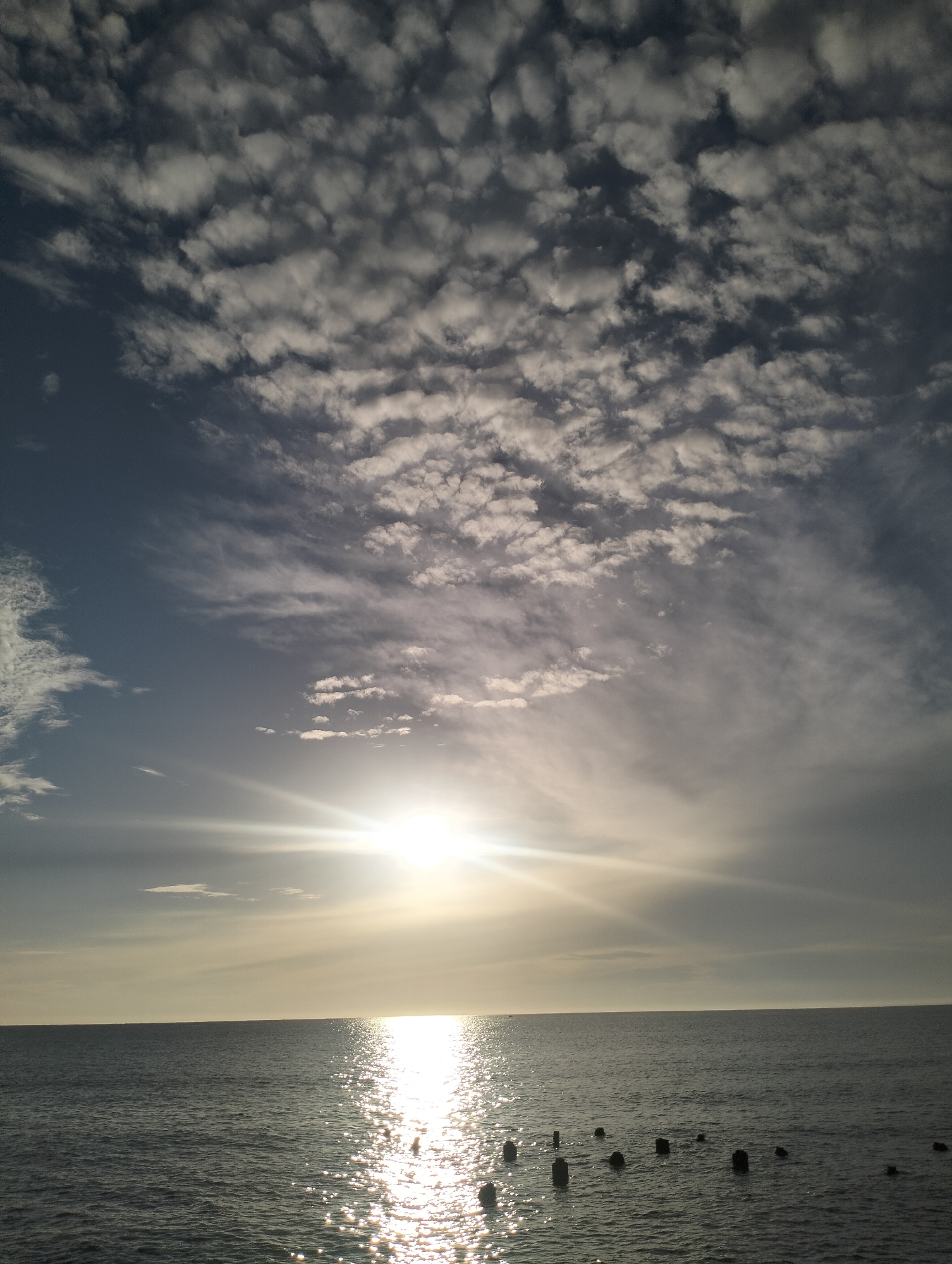 The image depicts a wide expanse of the sea under a dramatic sky filled with patchy clouds. The sun is low on the horizon, casting a bright glare across the water, creating a shimmering reflection. In the foreground, several dark, weathered posts or pilings protrude from the sea, scattered across the frame. The horizon line is clear, separating the dark blue water from the sky. The sky transitions from a bright, hazy area around the sun to a deeper blue towards the top. The overall impression is one of a serene, possibly late afternoon or early morning coastal scene. Based on the provided context, this scene is located in Lhokseumawe, Indonesia.