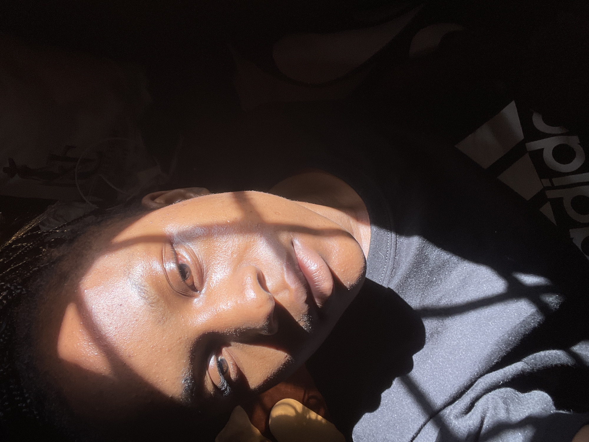 A medium shot captures a person's face illuminated by sunlight filtering through a window, creating a stark contrast between light and shadow. The person, facing the camera with a slight tilt of the head, has braids and appears to be wearing a black t-shirt with the Adidas logo visible on the chest. The lighting highlights the contours of their face, emphasizing their eyes, nose, and lips. The background is dark and indistinct, with a hint of shadows suggesting an indoor environment. The overall impression is one of dramatic lighting and a close-up portrait. The scene is located in Rafin Par, Nigeria.