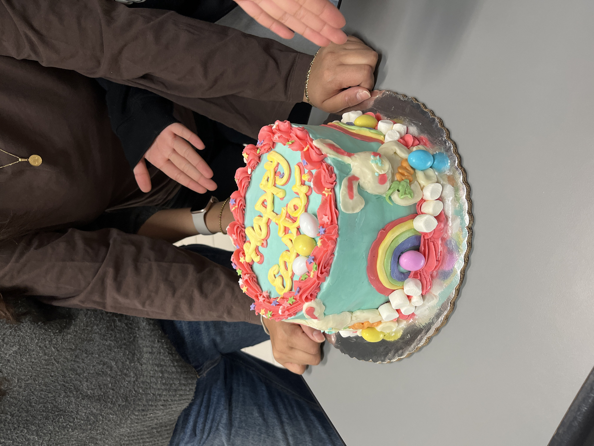 The image captures an indoor scene in Puebla, Mexico, focusing on a decorated cake placed on a light grey table. A round cake, adorned with light blue frosting, rests on a silver scalloped cake board in the foreground. The cake's top features pink icing rosettes bordering its edge, accented with colorful star and dot sprinkles. The phrase "Happy Easter" is piped in yellow icing across the center, accompanied by several white and yellow egg-shaped candies.
The side of the cake displays a white bunny figure with blue facial accents, holding an orange carrot. A rainbow arc in red, orange, yellow, green, and blue is depicted next to the bunny. White mini marshmallows and additional colorful egg-shaped candies in pink, blue, yellow, and orange are scattered around the base of the cake.
Several individuals are partially visible around the cake. On the left, a person wearing a grey knitted sweater and blue jeans has their right hand resting flat on the table. To the right, another person in a dark brown long-sleeved shirt, wearing a gold circular pendant necklace and a gold chain bracelet on their right wrist, has their left hand also resting on the table. A white smartwatch is visible on their left wrist. A third individual's hands are visible to the far right, hovering above the cake with palms down and fingers slightly spread. The background is indistinct and appears to be an interior space.