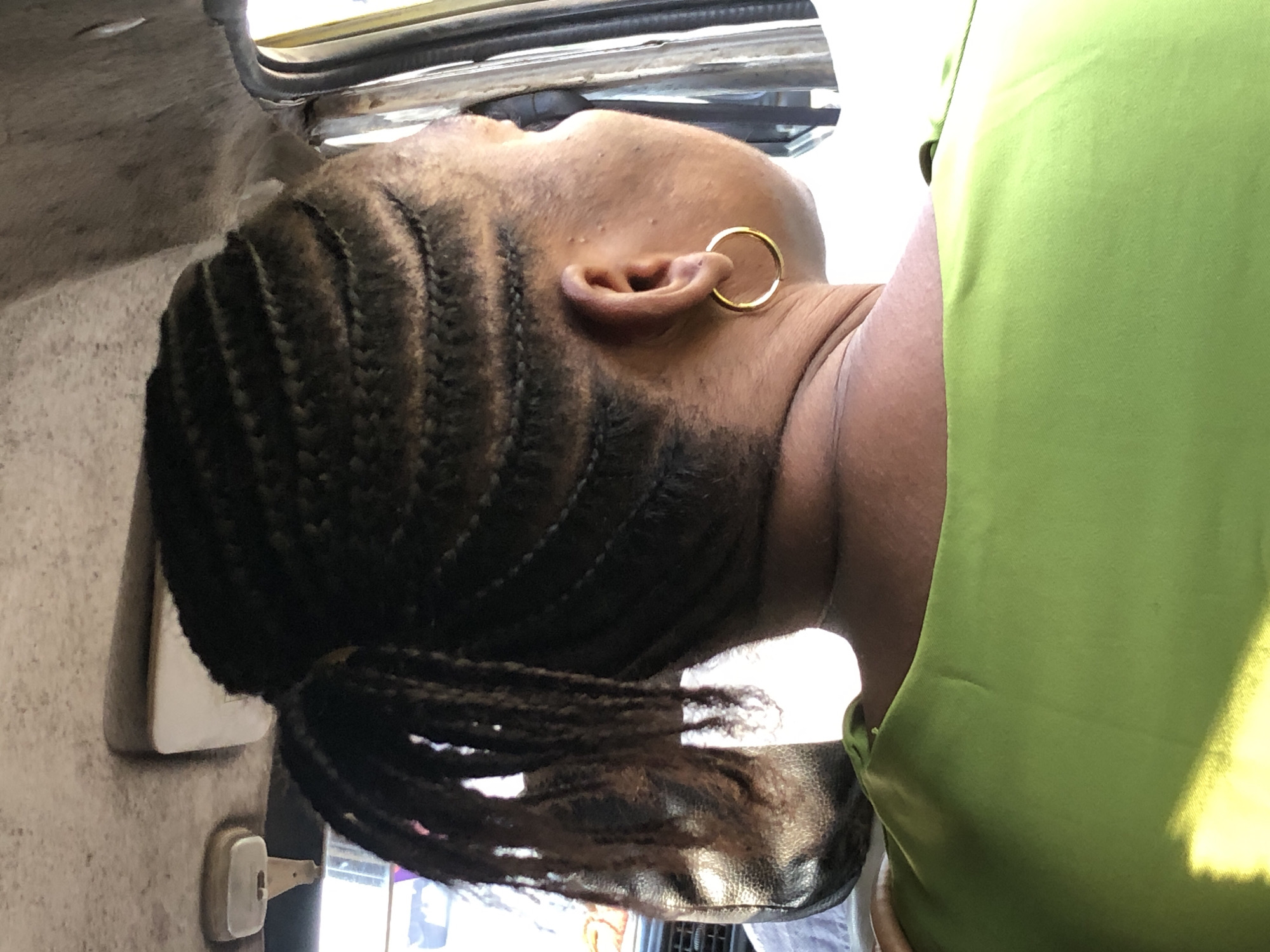 A medium-shot, over-the-shoulder, rear-view, outdoor portrait shows a Black woman with her hair braided into cornrows and a ponytail. She is wearing a lime-green top and a gold hoop earring in her right ear. The woman is inside a vehicle. The vehicle's interior includes a beige headliner with a plastic light fixture and a sun visor on the left side, and a black leather seat visible in the background on the left. The scene is brightly lit by sunlight. The image was taken on a sunny day in Ikeja, Nigeria.