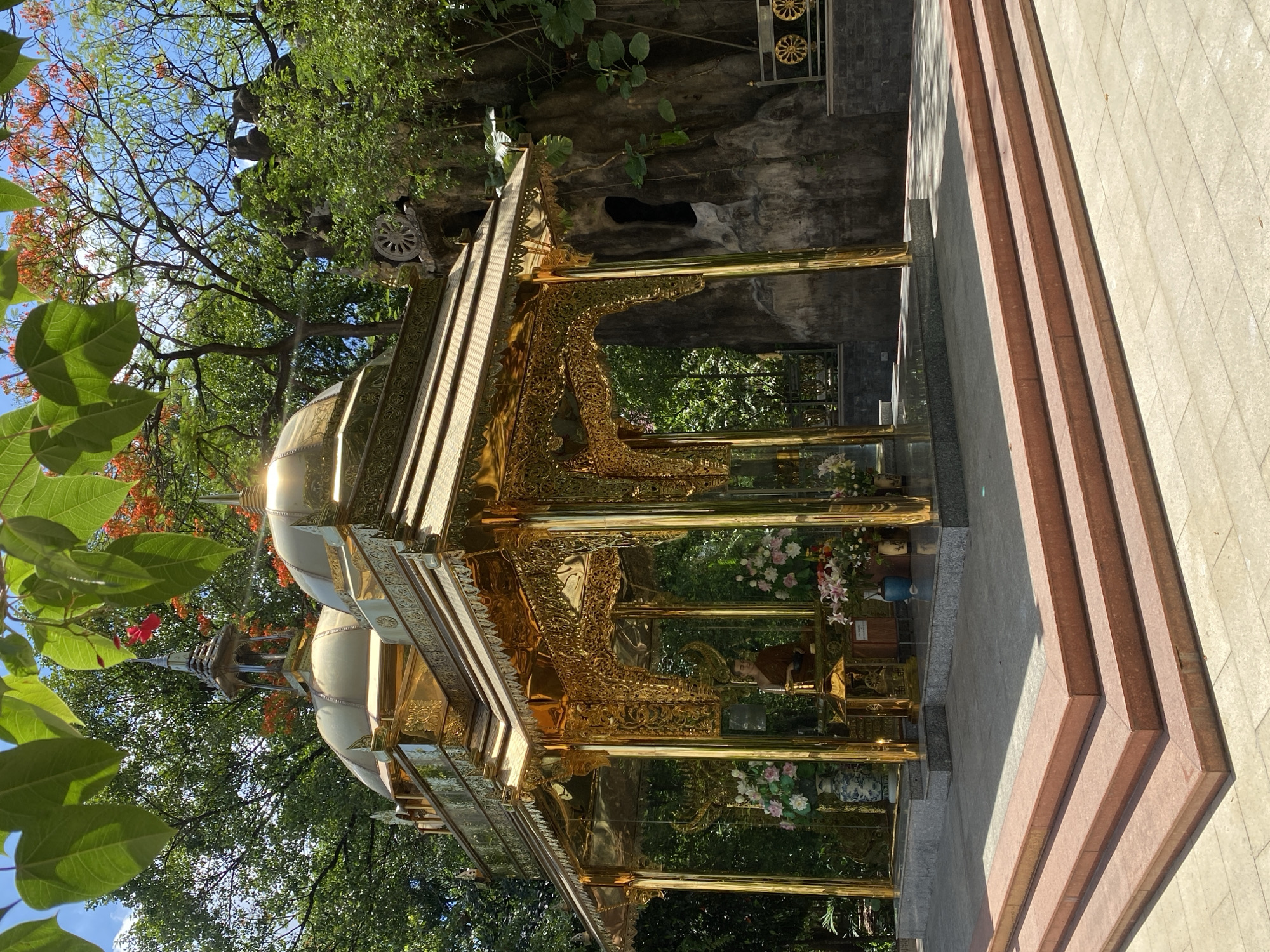 The image depicts a golden pagoda structure with a multi-tiered roof and intricate carvings, situated in an outdoor setting. The pagoda is partially obscured by foliage, with trees and flowering plants surrounding the structure. Steps lead up to the pagoda. The location is in Dĩ An, Vietnam.
