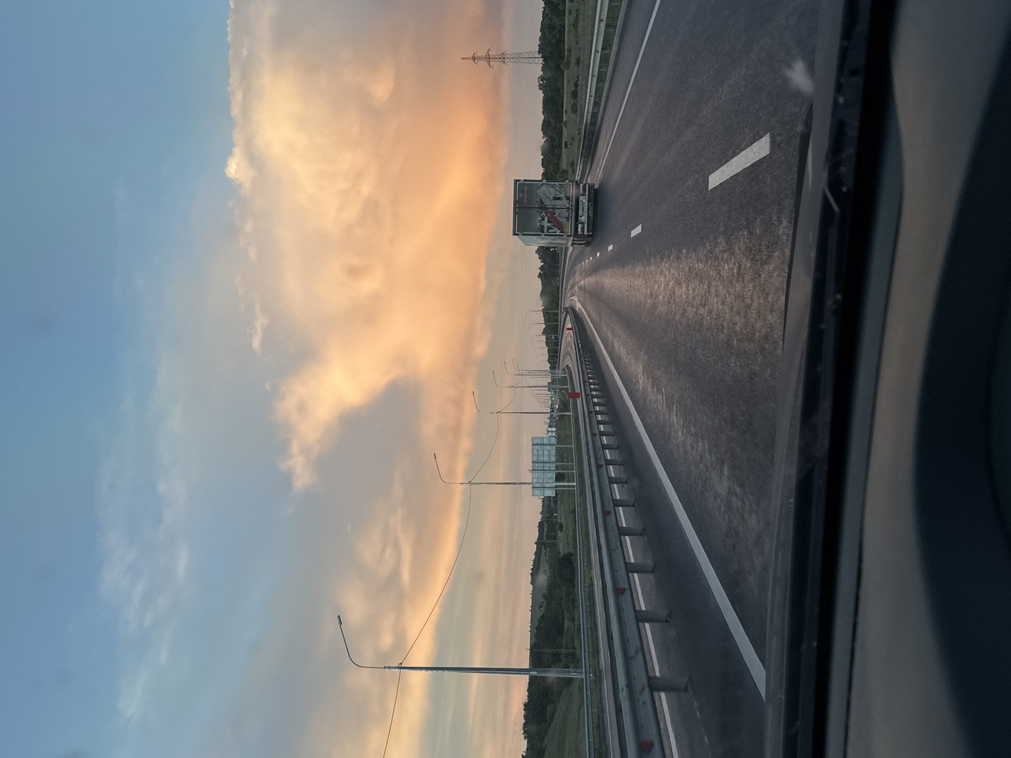 The image depicts a view from inside a moving vehicle on a highway in the Voskresensky District, Russia. The camera is facing forward, capturing the road ahead. A large truck is traveling in the same direction in front of the camera vehicle. The road is wet, and white lane markings are visible. A metal guardrail lines the side of the road. Beyond the guardrail, there is vegetation and street lights. The sky is cloudy with a sunset hue. An electrical tower is visible in the distance.