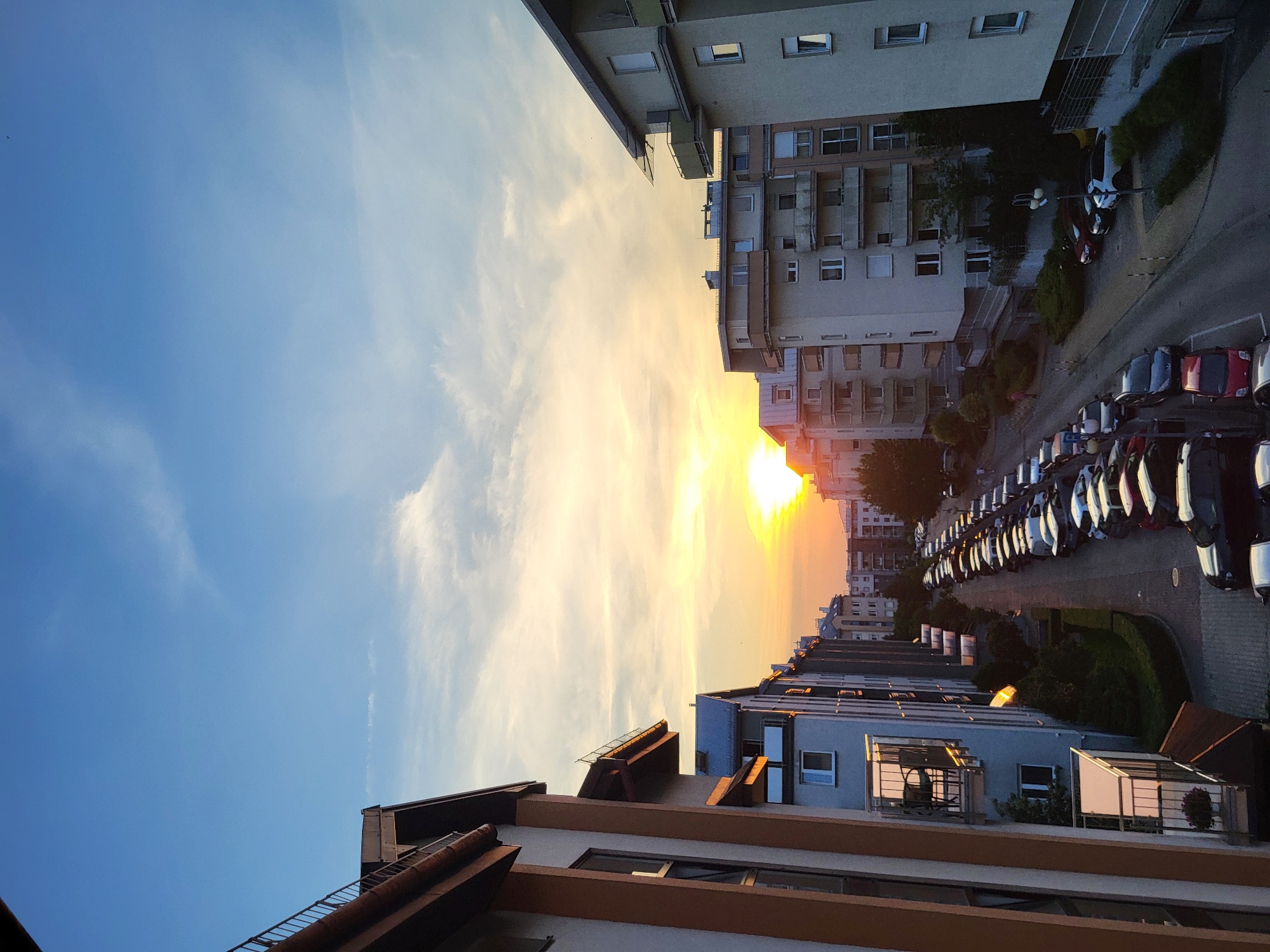 This is a vertical, eye-level outdoor photograph taken at sunset in a residential area of a city. The scene depicts a street lined with apartment buildings on both sides, filled with parked cars. The sky is a gradient of blue and orange hues, with wispy clouds illuminated by the setting sun. On the left side of the frame, an apartment building with beige and brown accents features balconies with railings and visible windows. The right side of the frame shows similar apartment buildings, also with balconies and windows. The street itself is paved and curves slightly to the right. The numerous cars parked along the street are of various makes and colors, mostly in a single lane. A few people can be seen walking on the sidewalk in the distance. The overall impression is that of a quiet urban evening.