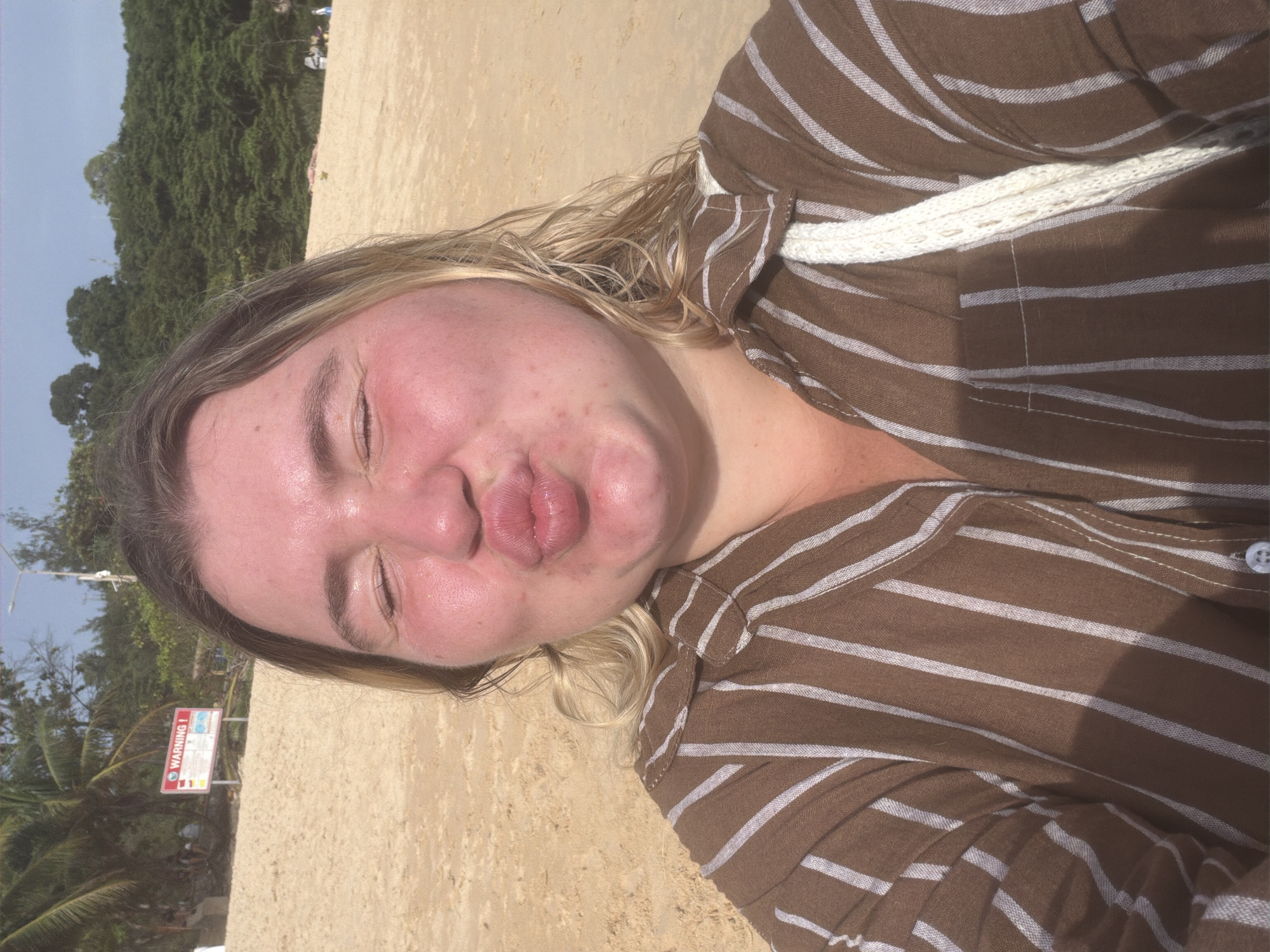 A woman with fair skin and blonde hair, wearing a brown and white striped shirt and a white woven bag strap, is taking a selfie on a beach. She is puckering her lips and has her eyes closed. Her face is flushed and appears shiny, possibly from sweat or sunscreen. In the background, there is a sandy beach, palm trees, lush green foliage, and a clear blue sky. A red and white warning sign is visible to the left, and there are other people and beach umbrellas further in the distance. The setting is indicative of a tropical beach location, consistent with Rawai, Thailand.