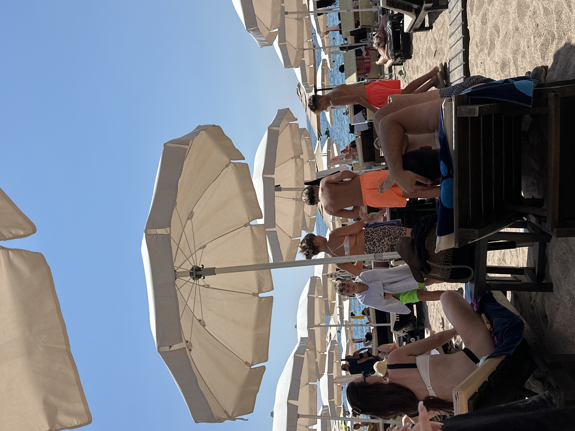 This is a wide shot of a crowded beach in Kamenovo, Montenegro, on a sunny day. Numerous beach umbrellas are set up along the sandy shore. In the foreground, several people are relaxing on lounge chairs. A woman in a white and black bikini is seated in a lounge chair facing left, with her legs extended. To her right, a man is lying on a lounge chair, with his arm raised to his head. In the mid-ground, a group of people are standing near the water. A young boy, wrapped in a white towel, stands facing the camera. Next to him, a young woman in a patterned bikini and leopard-print shorts is also facing the camera. Two shirtless young men in orange shorts are visible further back. The sea is visible in the background, with boats and people on the water. The beach is a mix of sand and wooden walkways.