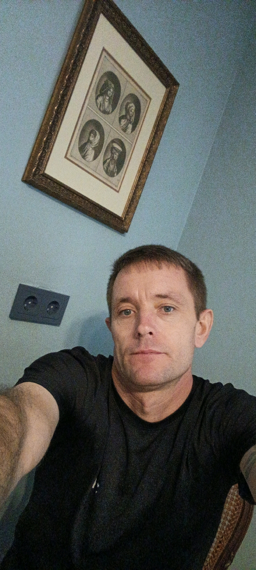 A man is taking a selfie in front of a pale blue wall. He is wearing a black t-shirt. Behind him, a framed picture hangs on the wall, containing four circular portraits. To the left of the framed picture, there is a dark rectangular electrical outlet. The man has short brown hair and blue eyes. He is looking directly at the camera. The overall scene appears to be indoors. There is no indication of this image being located in Tashkent, Uzbekistan.