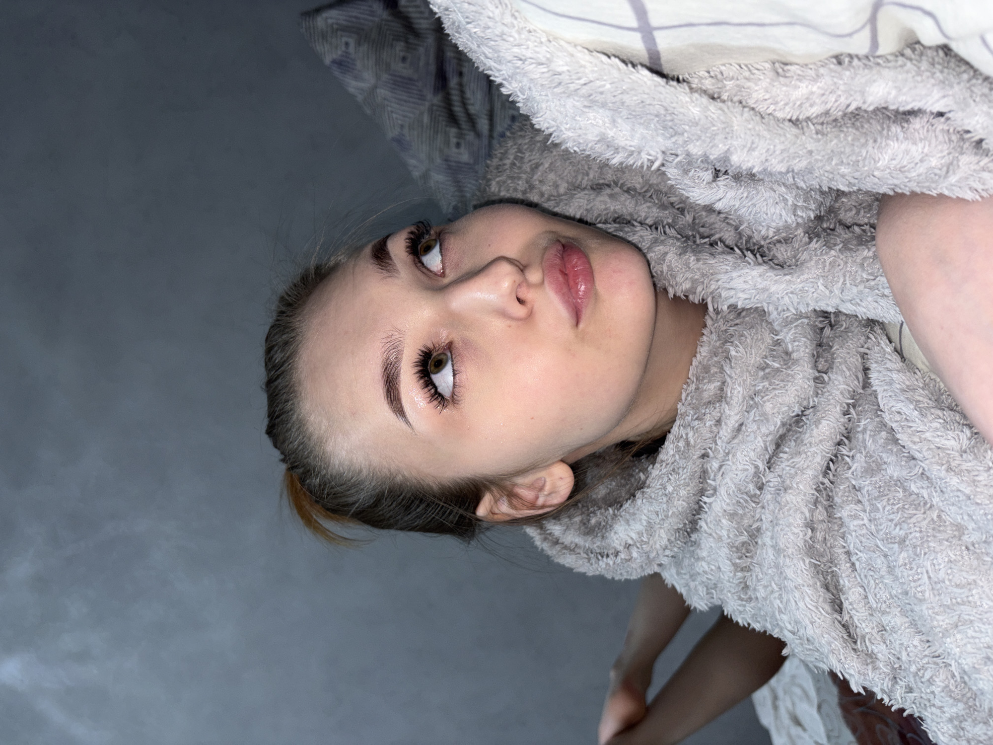 The image captures a close-up portrait of a young woman lying down indoors, viewed from an angled perspective with her head to the left. She is partially covered by a thick, plush, light grey blanket or robe, which frames her face and body. Her face is turned upwards, with her gaze directed towards the upper left, giving her a pensive or contemplative expression.
The woman has fair skin, prominent dark eyebrows, and long, dark eyelashes, which appear to be mascara-enhanced. Her eyes are a light brown or hazel color, and her lips are full and slightly parted. Her hair is dark at the roots with lighter strands and is pulled back, with a hint of a hair tie visible near her ear.
The background primarily consists of a plain, dark grey surface, likely a wall or floor, occupying the left side of the frame. To the upper right, a bed sheet or pillow with a subtle grey and white patterned design is visible beneath the fluffy blanket. Parts of her arm/hand and leg/knee are visible at the bottom edges, suggesting she is comfortably enveloped by the blanket.
The lighting is soft, creating gentle highlights on her face and the texture of the blanket. The overall atmosphere appears tranquil and intimate, suggesting a moment of relaxation. No specific time of day or weather can be discerned, but the setting is clearly indoors, possibly within a home in Chaikovsky, Russia, based on the provided location context. There is no visible text in the image.