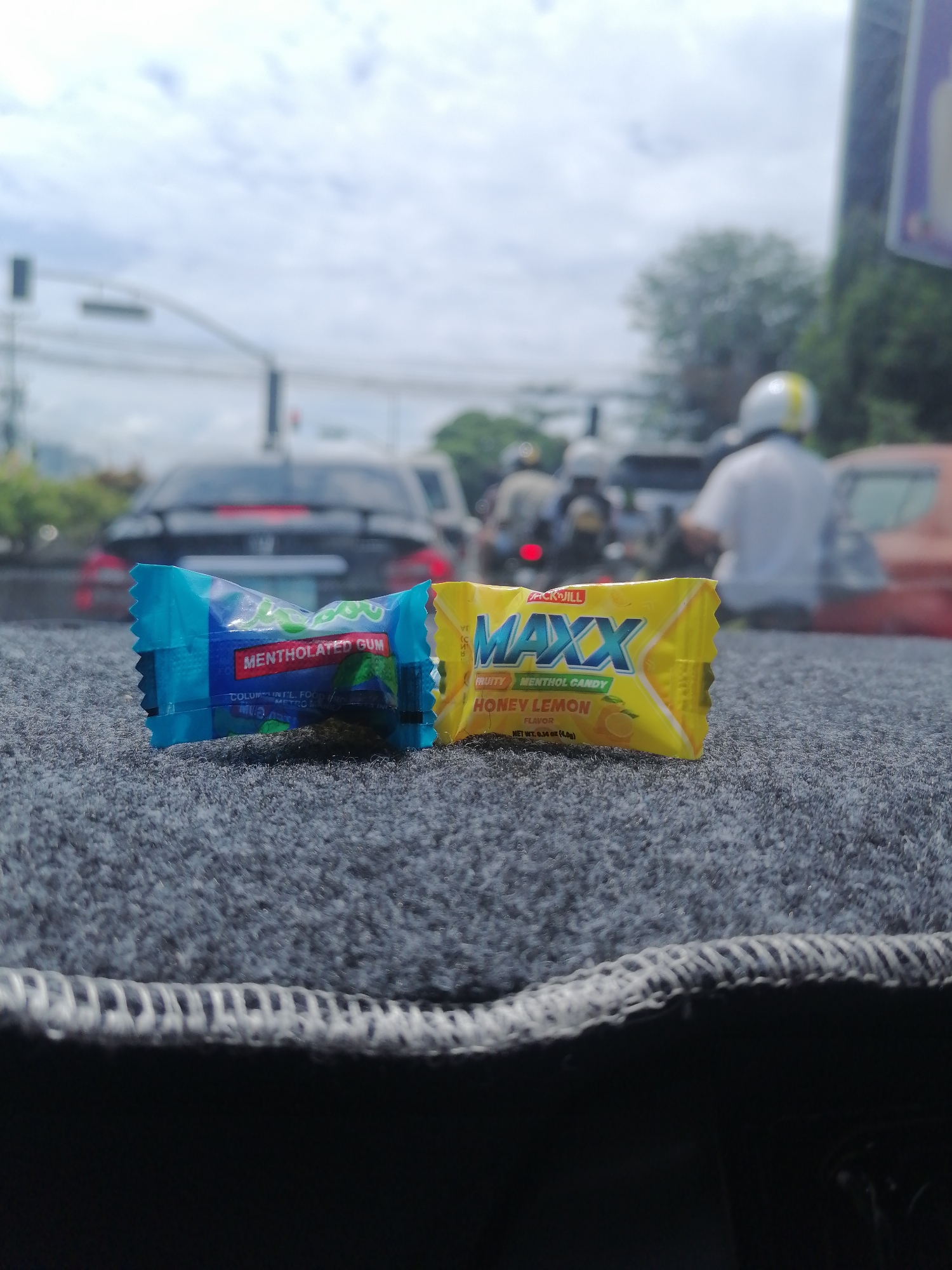 The image displays two individually wrapped candies resting on a textured, dark grey surface, likely a car's dashboard or floor mat. In the foreground, the focus is sharp on these items. The candy on the left is wrapped in blue packaging, visibly marked "MENTHOLATED GUM." Its brand name is partially obscured but appears to be "CLOROMINT." The candy on the right, in a bright yellow wrapper, clearly states "PICKJILL MAXX FRUITY MENTHOL CANDY HONEY LEMON FLAVOR" and "NET WT. 0.14 oz (4.0g)."
The background is heavily blurred, depicting a bustling street scene under an overcast sky, suggesting daytime. Traffic is visible, with several cars, including a dark sedan directly behind the candies and a red car to the right. Multiple motorcycles are also present, some with riders wearing helmets, one notably with a white helmet featuring a yellow stripe. A traffic light pole is faintly visible on the left. The overall setting points to an urban environment in Davao City, Philippines, characterized by the typical mix of vehicles and general street activity. The shallow depth of field ensures the candies are the primary subject, with the urban landscape serving as a soft, out-of-focus backdrop.