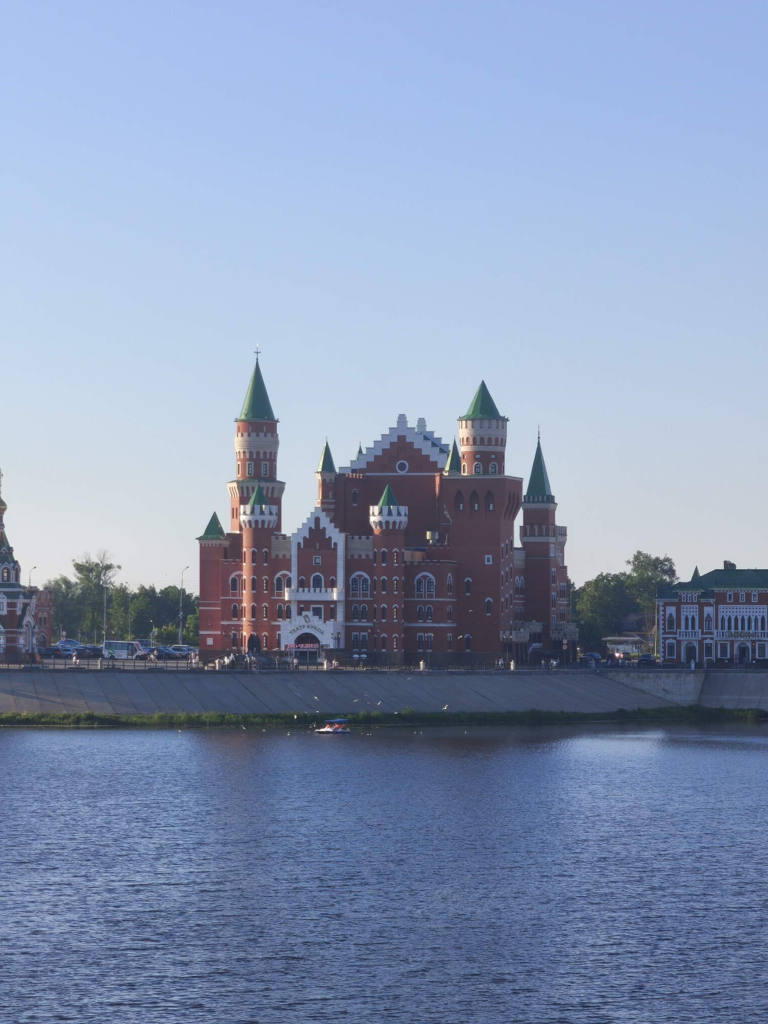 This is a full-shot, eye-level, medium-distance photograph of the National Puppet Theatre building in Yoshkar-Ola, Russia, as seen from across the Malaya Kokshaga River. The building, constructed in a neo-Gothic style with red brick and green spires, is the central focus. Several smaller, similarly styled buildings, including what appears to be a church to the far left, are visible to the left and right of the main structure.
In the foreground, the dark blue water of the river fills the bottom half of the frame, showing gentle ripples. In the middle ground, a concrete embankment lines the river, topped with a black metal fence. A paved walkway runs along the embankment, where several people can be seen walking. Cars and a white bus are parked or driving on the road behind the walkway. A small, light-colored boat with several occupants is visible on the water near the embankment, and numerous white birds are scattered across the water's surface and in the air above it.
The background is dominated by a clear, pale blue sky. The lighting suggests it is either early morning or late afternoon, with soft sunlight illuminating the scene. The overall impression is one of a well-maintained, picturesque urban waterfront.