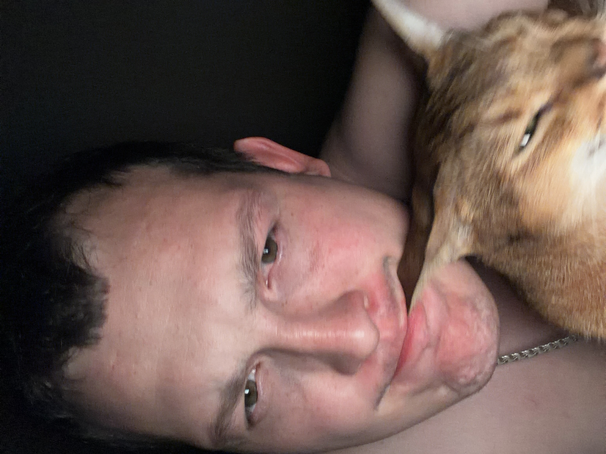This is a close-up, indoor, selfie-style photograph of a young adult male and a cat. The man's face is filling the left and central portion of the frame, and he is making a grimace or playful expression with his mouth. His right cheek is pressed against the cat's head. The cat, with orange and brown fur, is positioned on the right side of the frame, partially obscuring the man's face. The cat's head is turned towards the viewer, with its eyes partially closed. The lighting is dim, suggesting an indoor environment at night or in a dark room. The background is dark and out of focus. There are no discernible landmarks or objects that would indicate a specific location in the city of Chaikovsky, Russia.