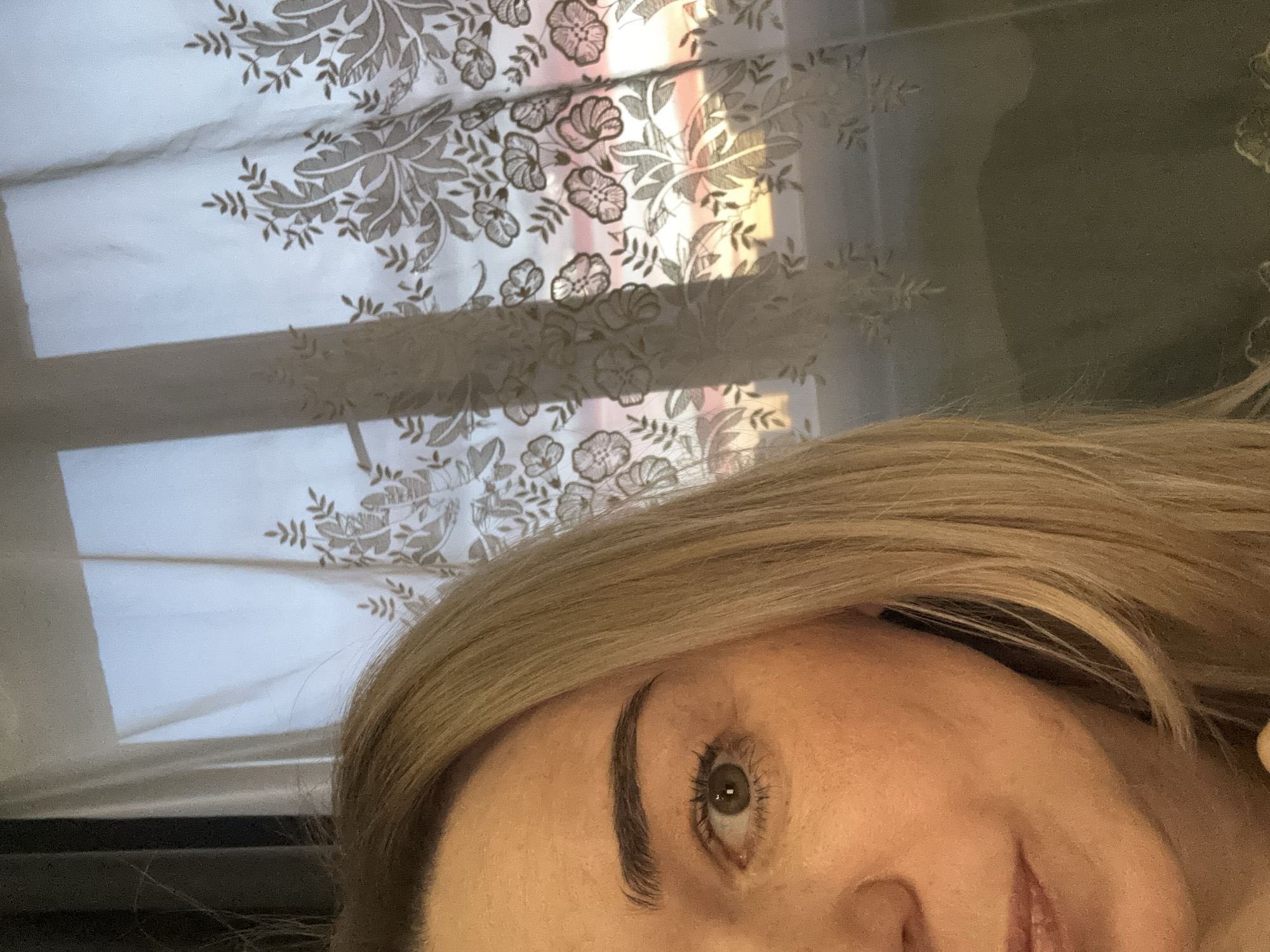 The image shows a partial view of a person's face, likely female, with blonde hair. One eye and eyebrow are visible, along with part of the nose and mouth. The background includes a window covered by a patterned sheer curtain. There is no information to indicate this image was taken in Ladozhskoe Ozero, Russia.
