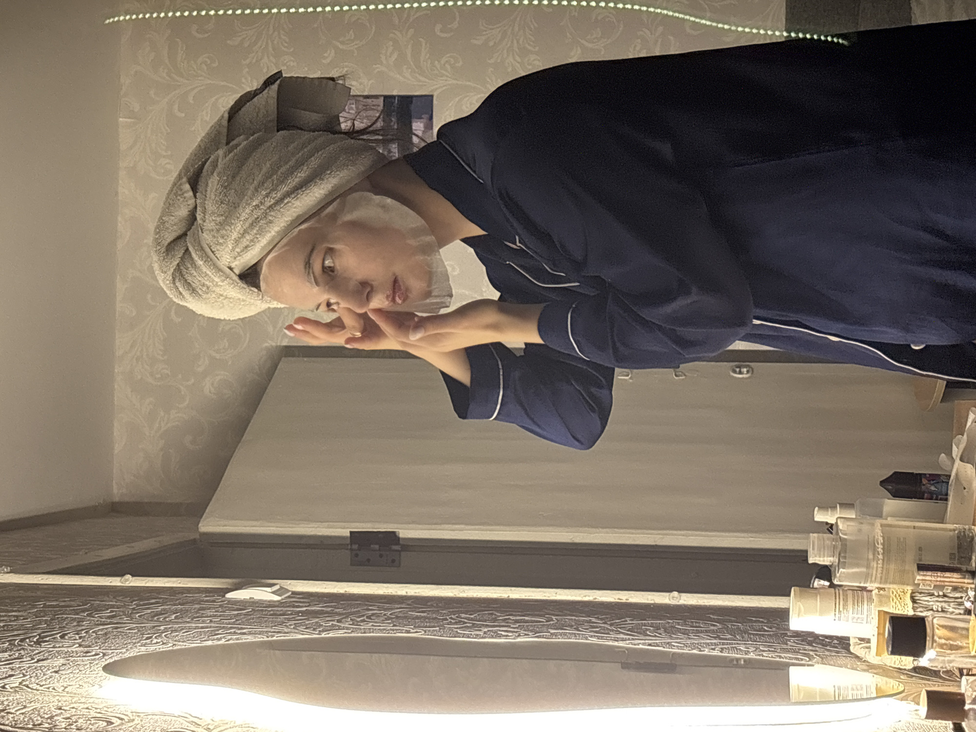 An indoor scene in Chaikovsky, Russia, shows a person engaged in a skincare routine during the evening or night. The person, likely female, is captured from the chest up, wearing dark blue pajamas with white piping and a light-colored towel wrapped turban-style around their head. They are applying a white product to their face with both hands, appearing focused.
The background features ornate, light-toned patterned wallpaper. Above, a strip of white LED lights illuminates the area. Below the person, reflecting in what appears to be a mirror, is a dark, patterned surface (likely a vanity countertop) on which numerous skincare bottles and jars are neatly arranged. A bright, oval-shaped light source is also visible in this reflection, further illuminating the counter and products. The overall lighting is artificial and gentle, suggesting a quiet, personal moment. No legible text is visible on any products or surfaces.