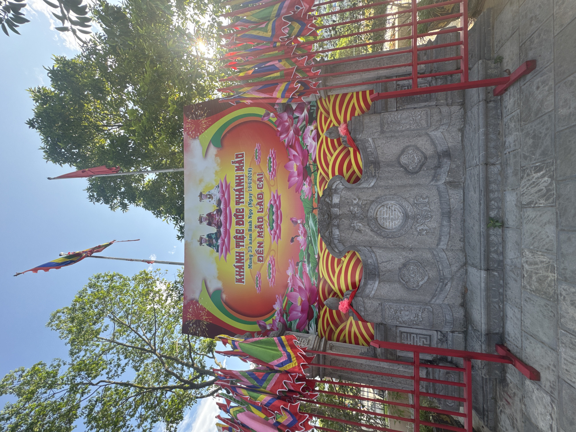 Here is a detailed description of the image:
The image captures a festive scene, likely a celebration related to "Đền Mẫu Lào Cai" (Lao Cai Mother Temple) in the city of Lao Cai, Vietnam.
The main focus is a large banner advertising a "KHÁNH TIỆC ĐỨC THÁNH MẪU" (Celebration of Mother Saint), which according to the print is to be held on "Mùng 3/3 năm Bính Ngọ (Ngày 19/4/2026)" (March 3rd, Binh Ngo Year, or April 19th, 2026). The banner is decorated with images of lotuses, a rainbow design, and what appears to be figures of deities. The banner is flanked by what looks like offerings or decorations in the form of red and yellow striped fabric.
The scene includes part of a stone structure, possibly part of the temple itself, with intricate carved details. A red metal fence surrounds the area. Colorful flags are placed along the fence.
The environment shows a partly sunny day with trees framing the scene. The flags and banner suggest a public and open setting.
