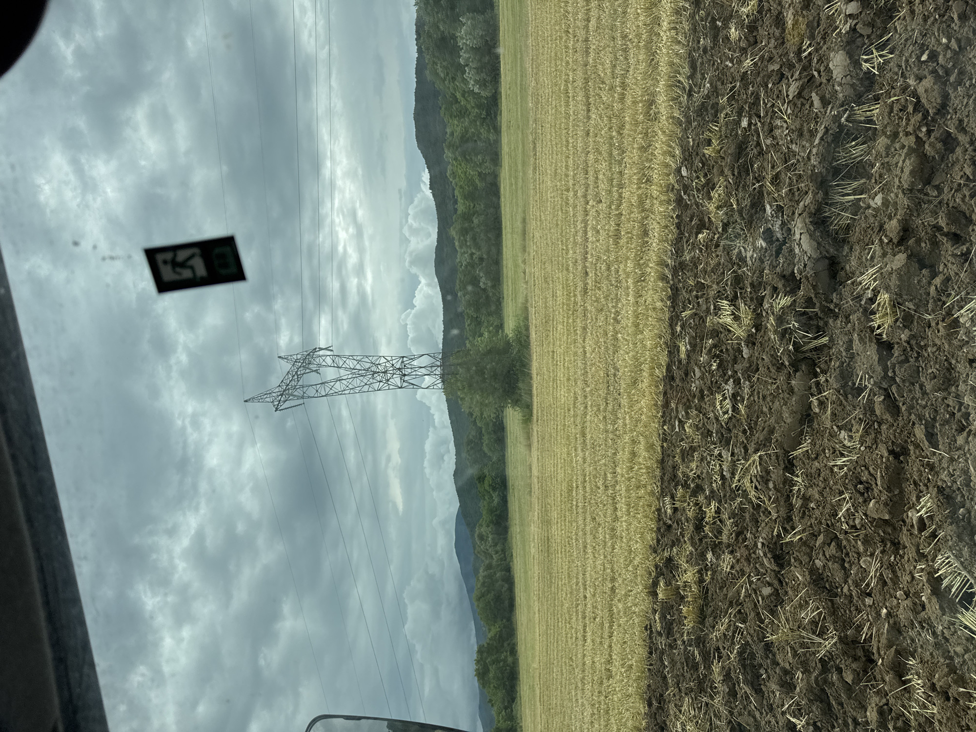 An overcast sky looms over a rural landscape in Prolom, Bulgaria. In the foreground, freshly tilled dark brown earth is visible, interspersed with dry straw. Behind this, a field of harvested golden crops stretches out, appearing as neat, parallel rows. A tall, latticed metal electricity pylon stands prominently in the middle ground, with power lines extending horizontally across the frame. A small, dark rectangular sign with a white pictogram of a running figure is hanging from the vehicle's interior, possibly an exit sign. The background features a dense line of green trees and rolling hills under a dramatic, cloudy sky.