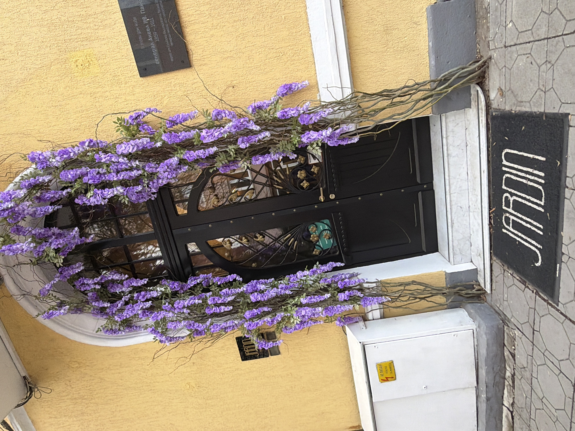 This image presents the exterior of "Jardin," a restaurant or similar establishment in Sofia, Bulgaria. The restaurant's entrance is decorated with cascading artificial purple flowers, framing the black metal door. A black welcome mat with "JARDIN" written vertically in white letters is positioned by the door. To the right, the building's exterior wall is clad in gray, textured tile, while the primary facade is painted yellow. There's a white electrical box and a commemorative plaque on the wall. The plaque, written in Bulgarian, provides historical information about the building. The environment around the entrance suggests a well-maintained urban setting.