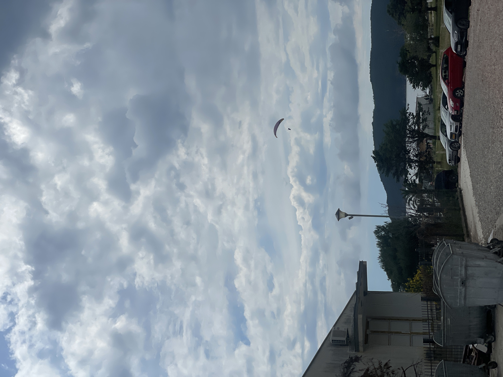 The photograph depicts a landscape scene in Boreti, Montenegro. The sky is predominantly cloudy with patches of blue. A paraglider is visible in the sky. In the mid-ground, there are trees and a mountain range. In the foreground, a row of parked cars line a road along with the corner of a building, a lamppost, and garbage receptacles.