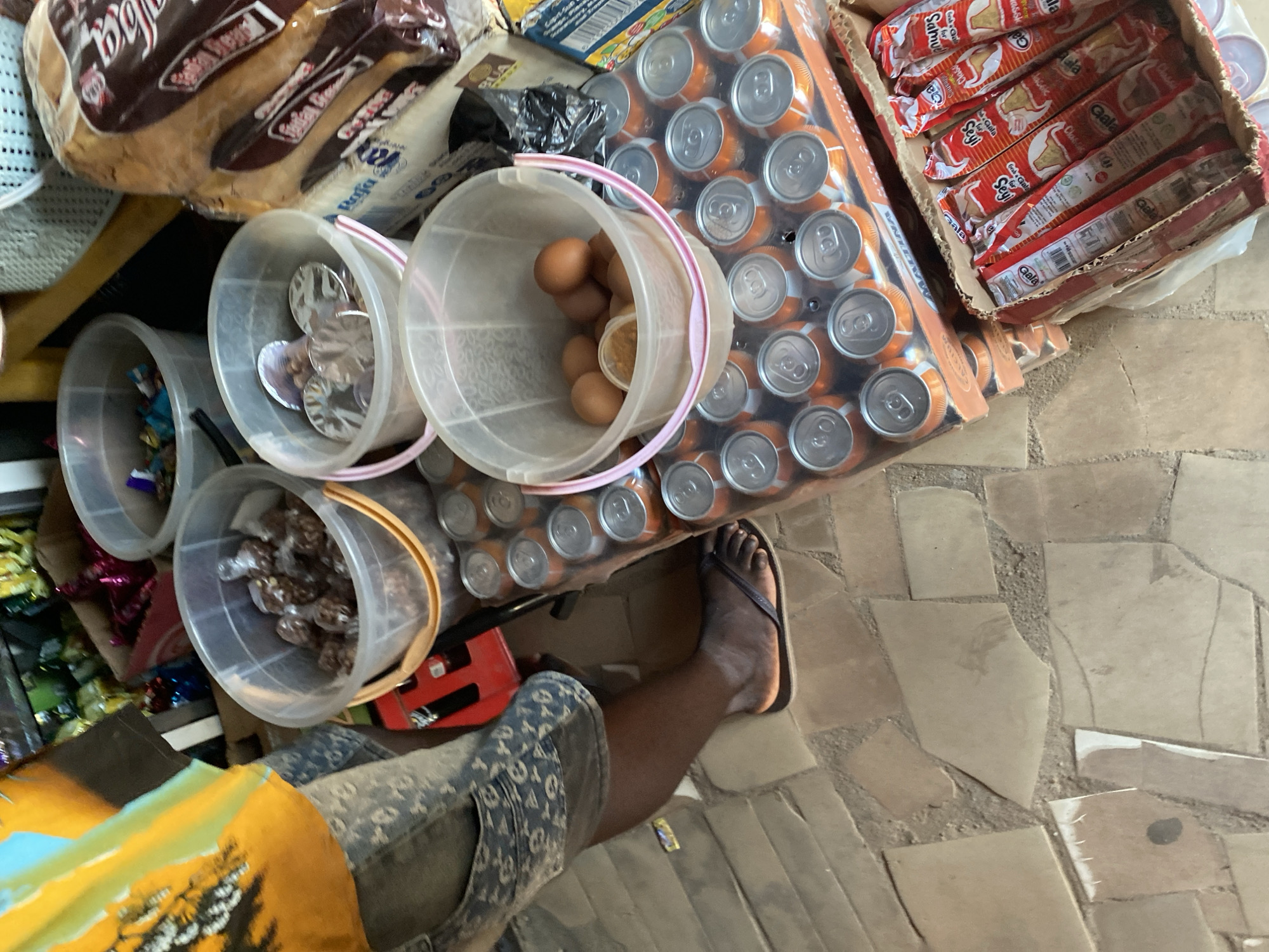 The image depicts a street vendor in Jalingo, Nigeria, standing behind a display of goods for sale. The vendor, partially visible, is wearing a patterned t-shirt and shorts. Their right leg is extended, and they are wearing dark flip-flops.
The foreground is dominated by a collection of beverages and food items. A stack of orange and silver soda cans is prominently displayed. To the right of the cans, a box contains several individually wrapped snack items, predominantly in red and white packaging with legible text.
Several transparent plastic buckets are also part of the display. One bucket contains what appear to be small, individually wrapped food items. Another bucket holds several brown eggs and a small container of a yellowish substance. A third bucket contains metallic discs, possibly lids or coasters.
The environment appears to be an outdoor market or street-side stall. The ground is paved with irregular stone tiles. In the background, more merchandise is visible, including bags of colorful snacks and other items, suggesting a bustling commercial setting. The lighting suggests it is daytime.