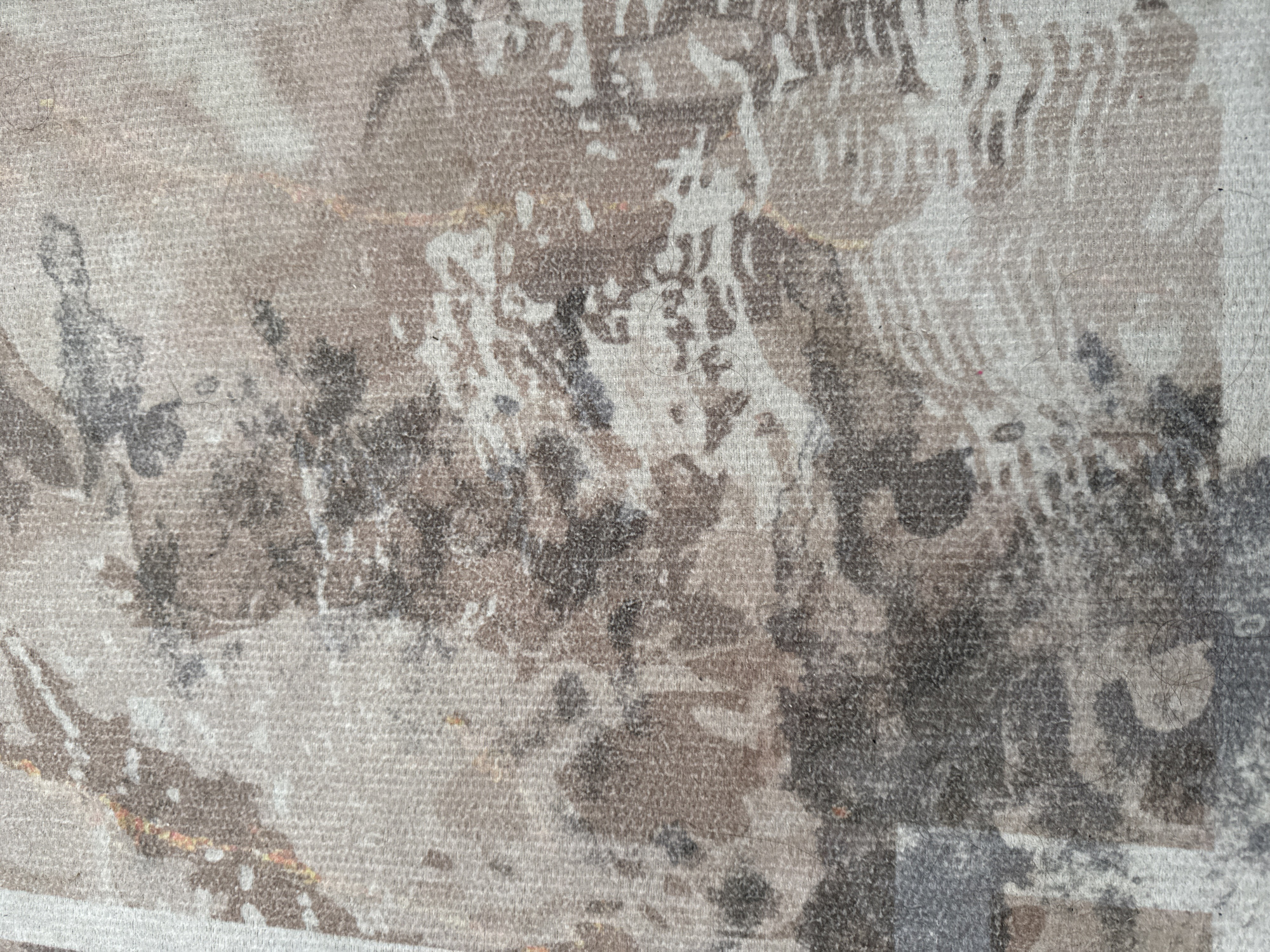 The image is a close-up, overhead shot of a textured carpet. The carpet has a distressed, abstract pattern with shades of beige, brown, grey, and some subtle hints of orange. The texture appears to be woven, with visible fibers and some loose strands of hair or lint on the surface. The pattern resembles watercolor splotches or marble veining, with areas of dark, irregular shapes against a lighter, textured background. There are no people, animals, or discernible objects in the frame, only the patterned surface of the carpet. The lighting is diffused, and the focus is sharp on the carpet's texture and pattern. The location in Gungoren, Turkey, is not visually evident from the image itself.