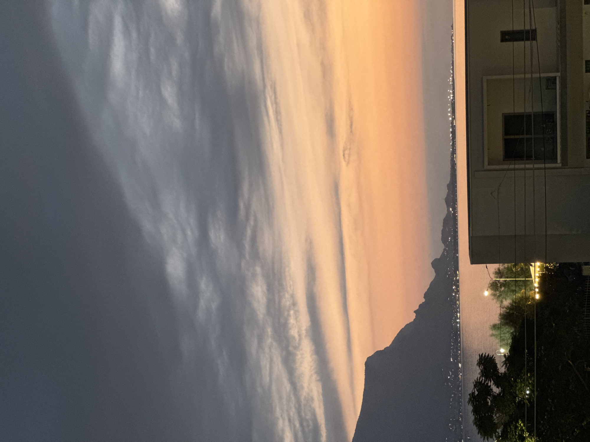 This image depicts a twilight scene in Capaci, Italy, characterized by a dramatic sky and a coastal landscape. The upper portion of the frame is dominated by dark, heavy clouds that transition into lighter, wispy streaks of gray and white, hinting at the retreating daylight. Below this, the sky is awash in hues of peach and orange, indicating the setting sun.
In the middle ground, a large, dark mountain range juts into the frame from the left, its silhouette sharply defined against the colorful sky. Along the base of the mountains, a line of lights suggests a coastal town or settlement. A body of water, likely the sea, lies between the mountains and the foreground, reflecting the soft light of the sky.
The foreground features a modern building with clean lines and a balcony. Electrical wires are visible in front of the building, adding a man-made element to the natural scenery. To the left of the building, dark foliage and trees are present, interspersed with the warm glow of streetlights and other artificial illumination, suggesting the presence of urban development or a well-lit area. The overall impression is one of a serene, yet atmospheric, evening view of the coast. There are no people or specific actions depicted in the image.
