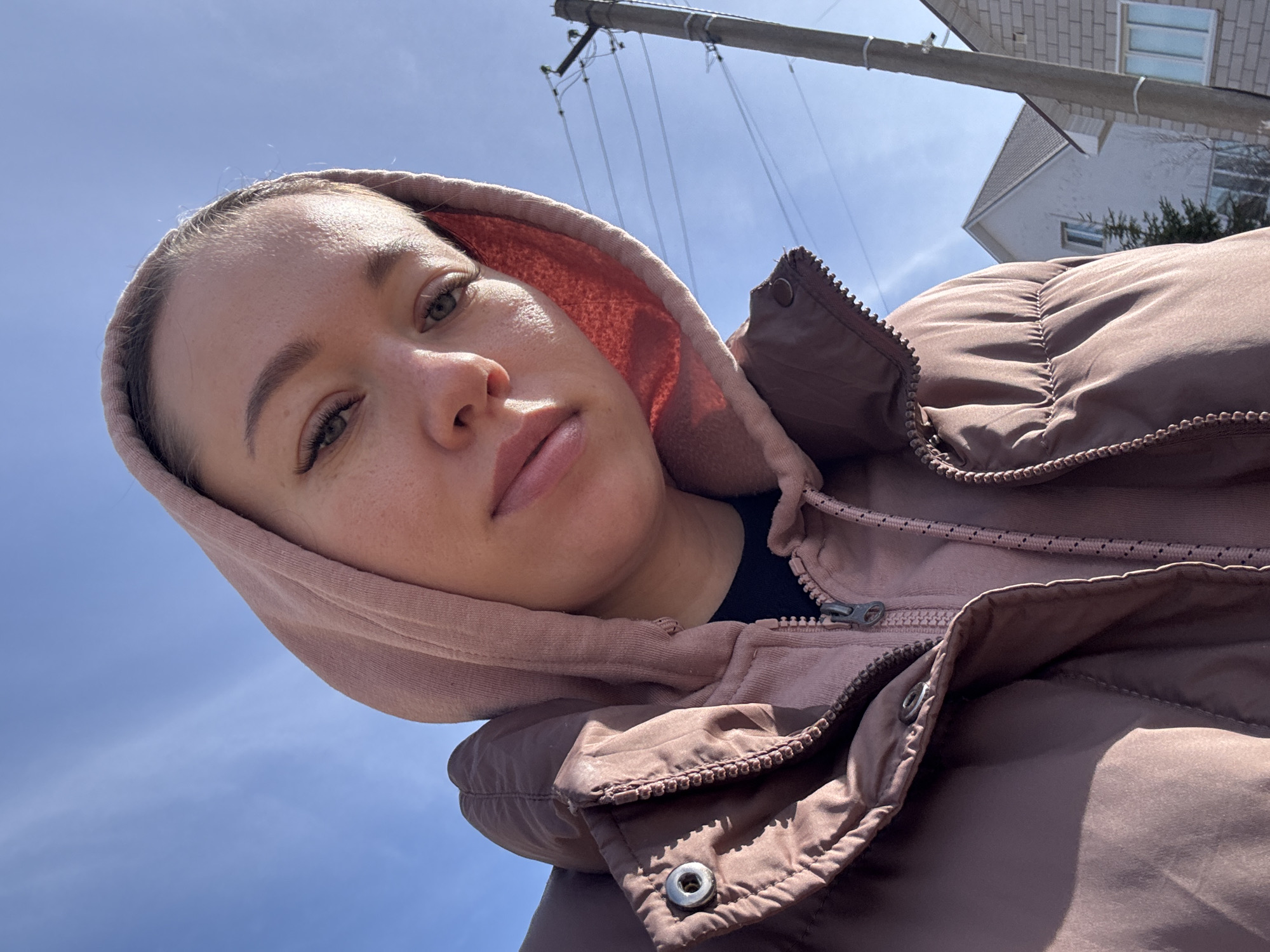 A low-angle shot depicts a woman wearing a brown winter coat with a pink hoodie, her face turned towards the camera. The background reveals a blue sky, a utility pole with wires, and a building constructed from light-colored bricks. The scene appears to be located outdoors, possibly in a residential area in or near the city of Ferma, Russia.