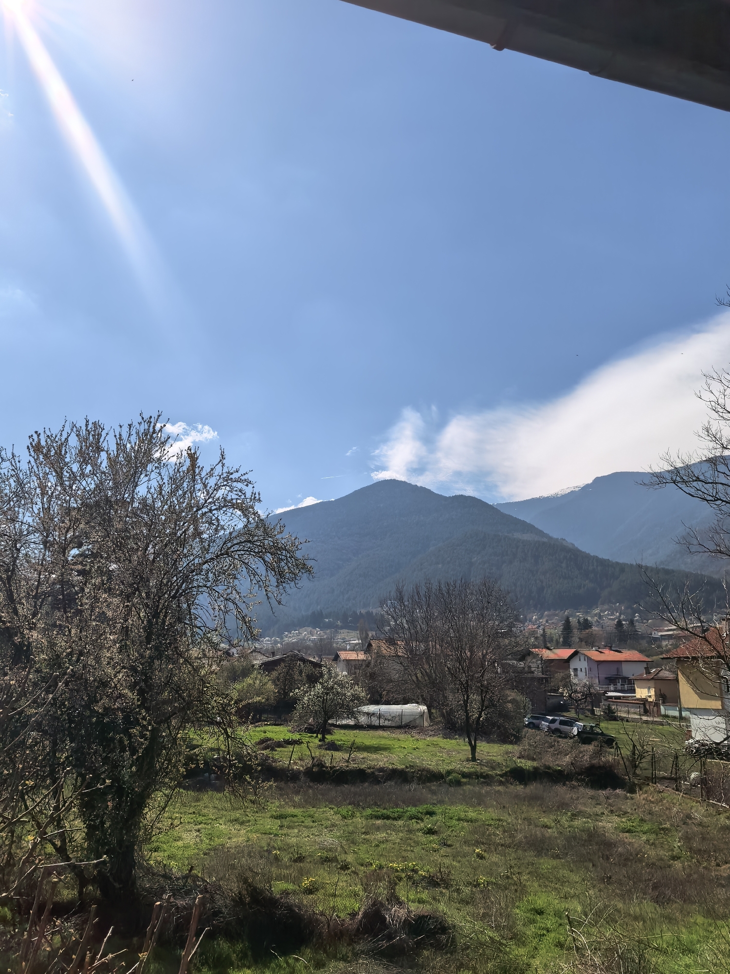 The image depicts a landscape scene in Unknown, Unknown, featuring a green field in the foreground with scattered trees, some still bare and others starting to bloom. Behind the field, there's a cluster of houses with orange roofs, suggesting a residential area. Further back, a range of mountains rises, some covered in dense forest and others showing rocky outcrops. The sky is mostly blue with some white clouds and a strong sun glare in the upper left corner.