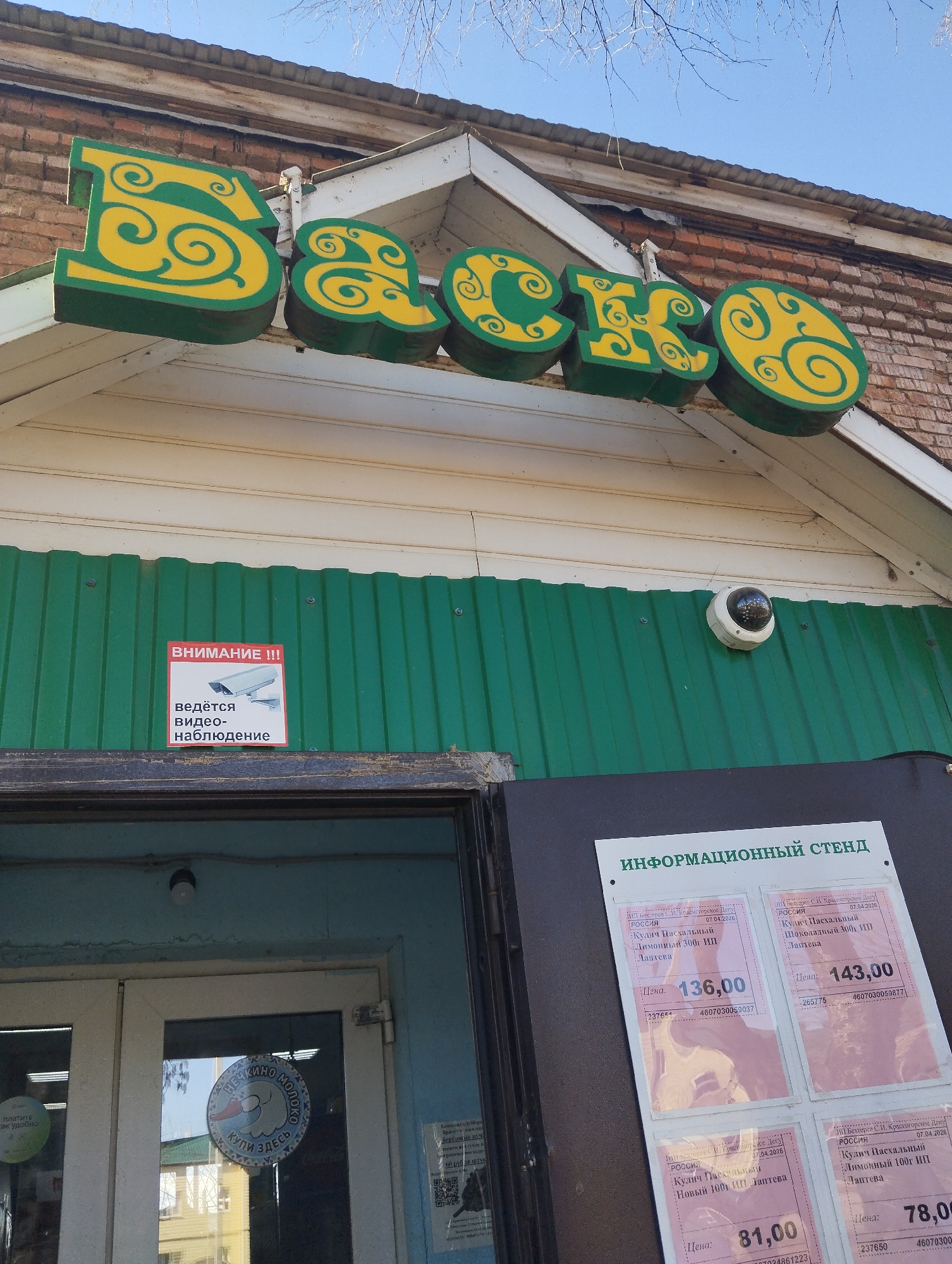 The image shows the exterior of a building, likely a store, in Krasnogorskoye, Russia. Above the entrance is a green and yellow sign with the name "Васко" (Vasco). A security camera is mounted above the doorway. A sign with the text "ВНИМАНИЕ !!! ведётся видео-наблюдение" (WARNING !!! video surveillance in progress) is posted to the left of the camera. To the right of the doorway is an information board with price lists for "Кулич Пасхальный" (Easter cake) items, including variations like "Лимонный" (Lemon) and "Шоколадный" (Chocolate), from ИП Лаптева (IP Lapteva, a private entrepreneur). The prices are in rubles, and the dates on the information are April 7, 2026. A sticker on the door has the text "Нечкино Молоко. Купи Здесь" (Nechkino Milk. Buy here).