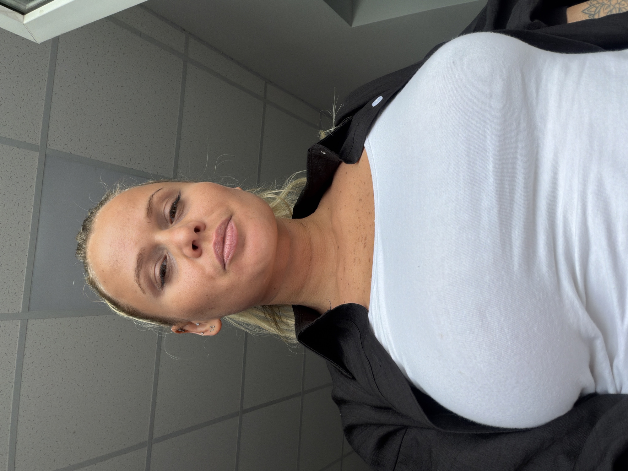 A low-angle, medium shot shows a blonde woman in a black open shirt over a white tank top, with her head tilted back slightly as she looks at the camera. Her eyes are closed, and her lips are parted. The woman has light skin with freckles and is wearing small stud earrings. The background appears to be an indoor office space with a textured ceiling and a white window frame visible on the right. The lighting suggests an overhead fluorescent light source.
