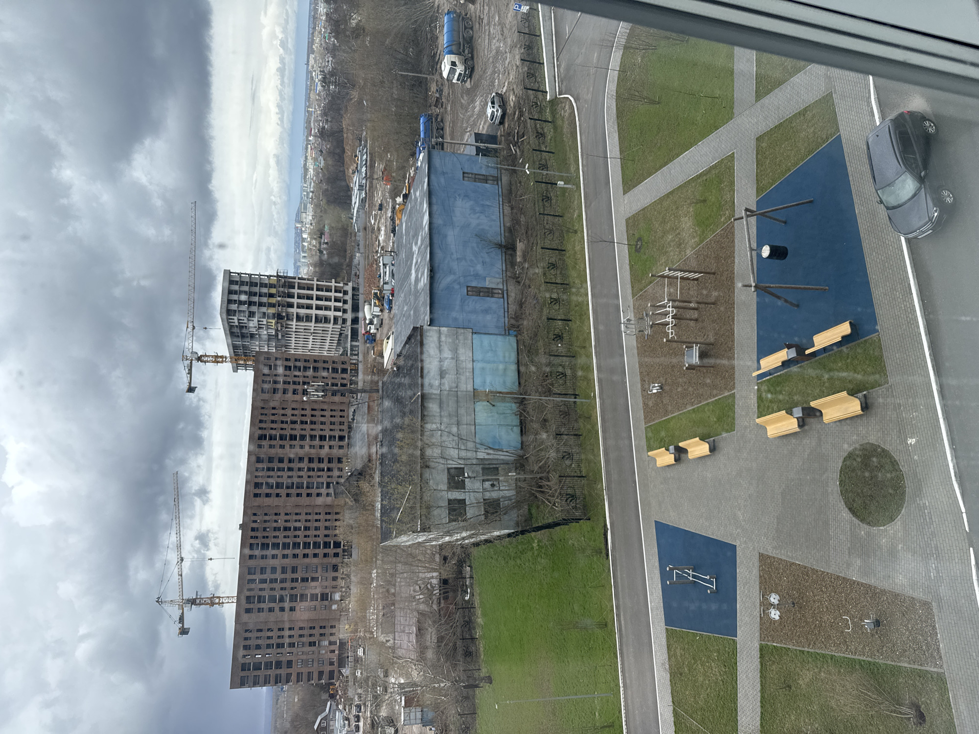This high-angle, panoramic view from a building overlooks an urban landscape in Matiushino, Russia, under an overcast sky.
In the immediate foreground, a modern recreational area is visible, featuring paved walkways, patches of green grass, and designated play or exercise zones with blue rubberized surfaces and sand-colored ground cover. These zones contain various equipment, including monkey bars, a small balance beam, and other fitness structures. Several light-colored wooden benches are placed along the paved paths. A dark grey car is parked on a paved road bordering this area.
Further into the midground, a large, rectangular industrial-style building with blue siding stands. Adjacent to it is an active construction site with several large trucks, including cement mixers, and piles of earth or materials. Scattered bare trees indicate late winter or early spring. A blue "P" sign with an arrow, indicating parking, is visible on a post near the construction vehicles.
The background features multiple tall, multi-story buildings. Some appear to be completed residential structures with numerous windows, while others are clearly under construction, showing concrete frameworks and scaffolding. Several prominent yellow tower cranes are visible, reaching high into the heavy, dark grey cloud-filled sky. A distant cityscape or town is discernible on the horizon, blending with the cloudy weather conditions.
No people are visible engaged in activities within the scene. The overall impression is one of ongoing urban development, contrasting newly completed public spaces with active construction and industrial elements.