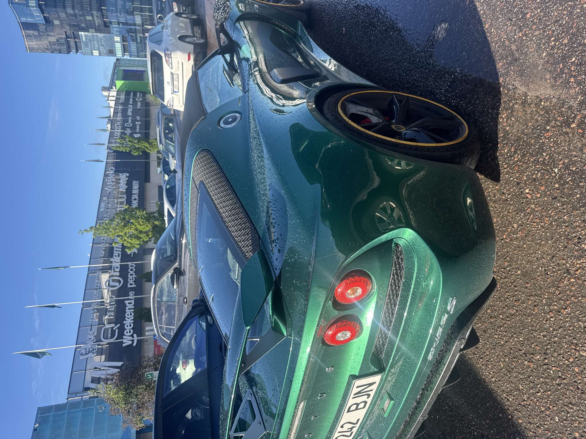 A green Lotus Evora is parked in a wet asphalt parking lot in front of a large commercial building with multiple storefronts and flags. The building has signs that read "Weekend.ee" and "Pepco". In the background, a modern glass skyscraper is visible. The sky is blue with scattered clouds. The car has a license plate that reads "242 BJN". The car is wet, indicating it may have recently rained. There are no people visible in the image.
