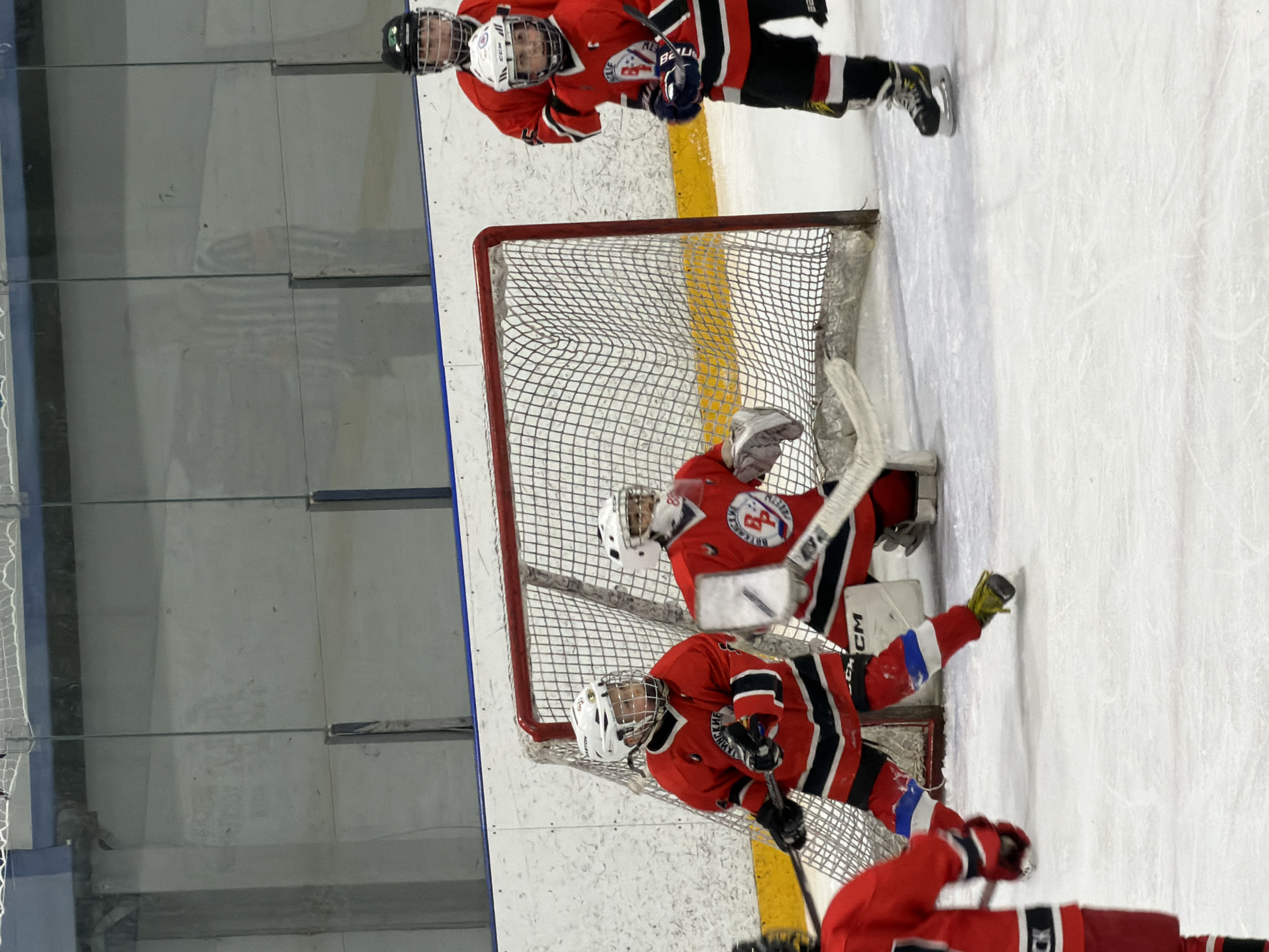 An indoor ice hockey rink in Votkinsk, Russia, is captured during what appears to be a youth game or practice. The image features at least five young hockey players, all wearing matching red jerseys with black and white stripes, black pants, and various helmets.
In the center, a red-framed hockey net with white mesh stands prominently. A goaltender, clad in a red jersey, white helmet, and full white pads, blocker, and catcher, is positioned behind the net, crouched and ready. The brand "CM" (likely CCM) is visible on the goalie's leg pads. Another player in a red jersey, wearing a white helmet with "CCM" branding, kneels in front of the net, holding a hockey stick.
Further back, two additional players in similar red jerseys and black helmets are visible near the top of the frame. One of the jerseys clearly displays "BOTKINSK", indicating the team or location, along with a circular logo. A player in the bottom right foreground is partially visible, also in a red jersey. Hockey sticks, some bearing the "BAUER" brand, are held by the players.
The ice surface is white, marked with a yellow line near the net. Clear glass panels line the perimeter of the rink, typical for an indoor hockey venue. The overall scene suggests an active and focused moment during the hockey session.