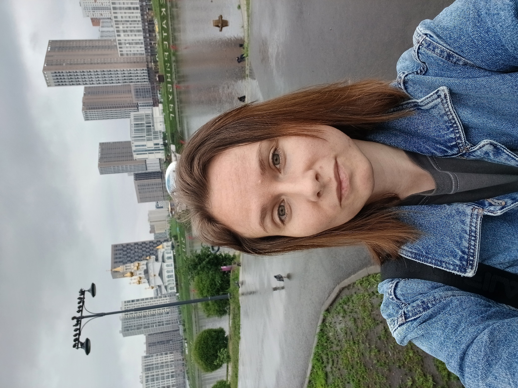 The image is a selfie taken by a woman in Yekaterinburg, Russia, near the Iset River embankment. In the foreground, the woman is wearing a denim jacket and a grey t-shirt. Her face is visible, with brown hair. In the background, a cityscape is visible with modern high-rise buildings and a church with golden domes. The ground is wet, suggesting it has recently rained. A street lamp with several birds perched on it is also visible, along with a few pigeons on the wet pavement. A sign with Russian text, appearing to be "ЕКАТЕРИНБУРГ" (Yekaterinburg), is reflected in the water.