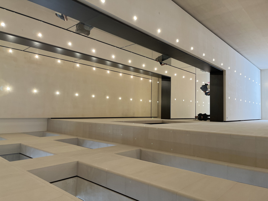 The image depicts the interior atrium of the Apple Store Champs-Élysées, located at 114 Avenue des Champs-Élysées, Paris, France. The perspective is a low-angle shot looking upwards within a modern, multi-story architectural space.
On the left, a tall, cream-colored stone wall extends vertically, featuring multiple recessed rectangular window openings. On the right, several tiered floors or balconies project into the atrium, each defined by dark, linear railings with glass panels. The ceilings of these levels are light-colored and uniformly illuminated by numerous circular recessed spotlights.
Observations of individuals include:
* On an upper balcony, a person is partially visible, leaning over the dark railing, seen from behind.
* On a lower balcony, a security guard in a dark uniform stands near a light-colored column on the left side.
* Further along the same lower balcony, two other individuals are positioned near the railing, appearing to be seated or crouched.
The overall environment is brightly lit, spacious, and characterized by a minimalist, contemporary design with clean lines and reflective surfaces.