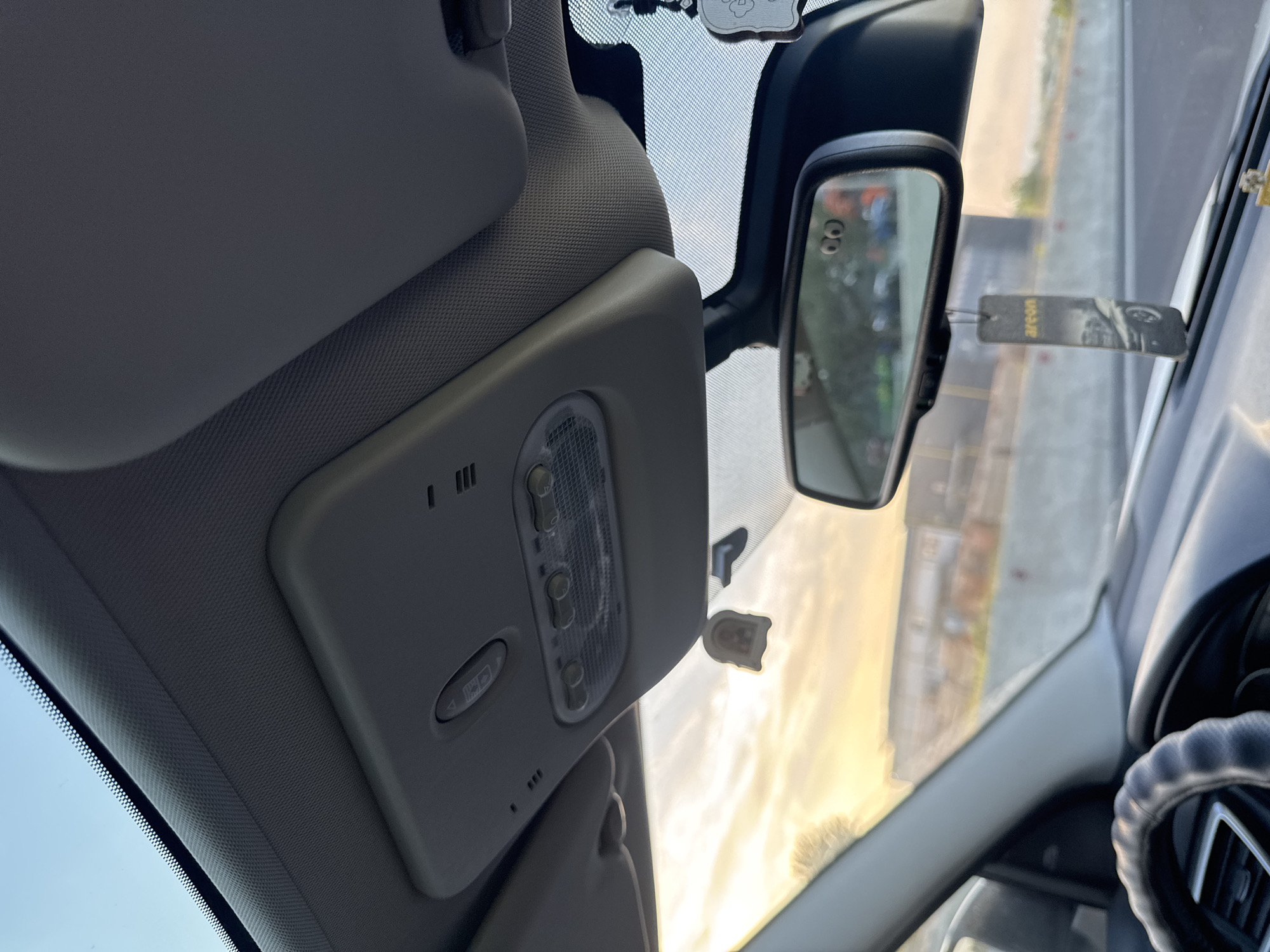 The image shows the interior of a vehicle, looking forward through the windshield. The sky is visible with clouds and a sunset. In the foreground, a steering wheel is partially visible. Above the windshield, there is a console with buttons and lights, and a rearview mirror. A car air freshener hangs from the rearview mirror. The background appears to be a highway with some buildings. There are no people visible in the image. The location is identified as Cojușna, Moldova.