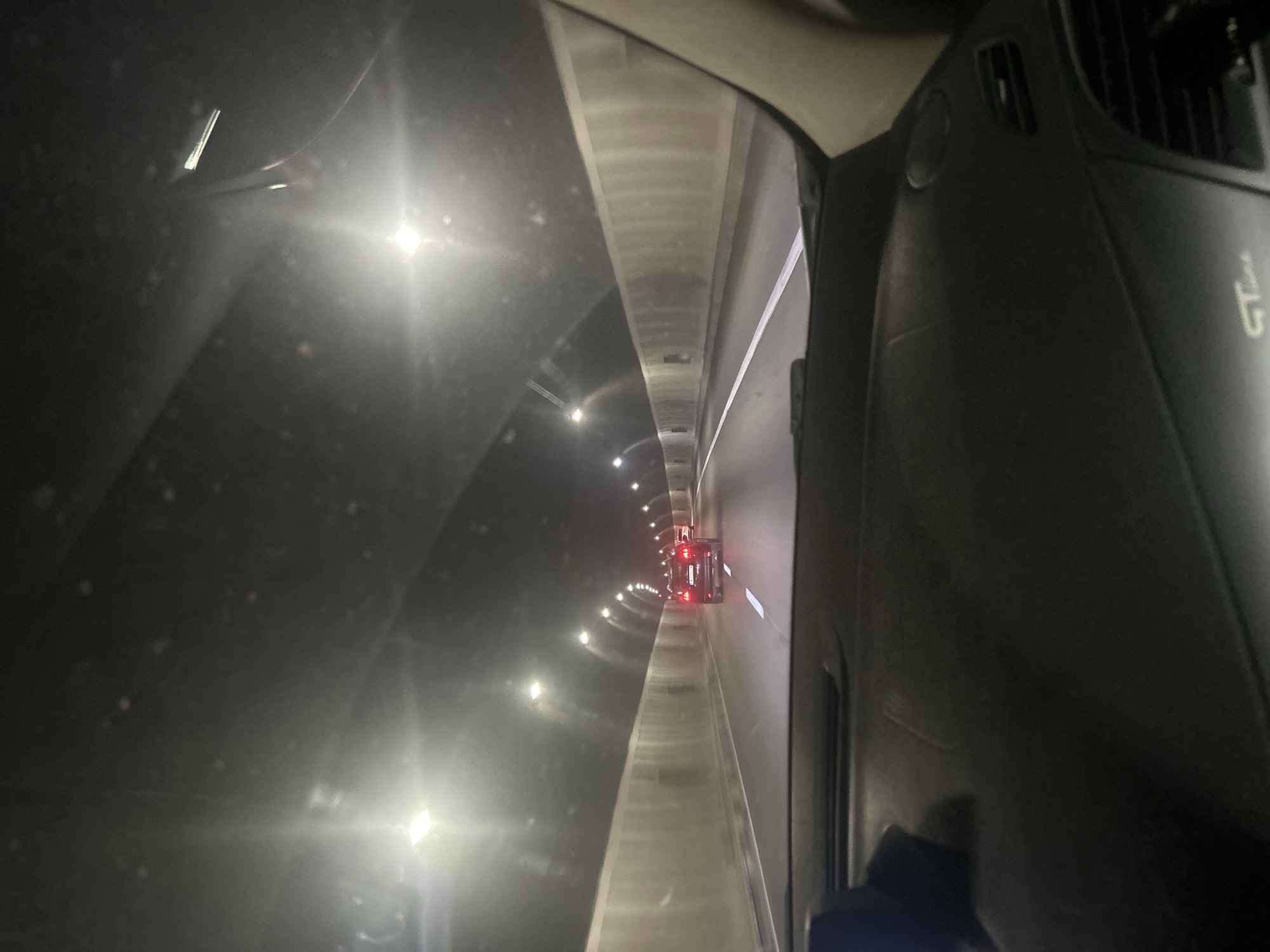This media file captures a view from inside a vehicle traveling through a tunnel in Dolna Vasilitsa, Bulgaria. The scene is dark, illuminated by the tunnel's lighting system, which consists of a series of lights running along the ceiling and walls. The tunnel appears to be a road tunnel, with a lane visible ahead leading into the distance. A red vehicle with illuminated taillights is visible further down the tunnel, suggesting other traffic is present.
The perspective suggests the photo was taken from the driver's or passenger's seat, looking forward through the windshield and side window. Part of the vehicle's interior, including the dashboard and a vent, is visible on the right side of the frame, indicating the point of origin of the photograph. The tunnel's architecture is somewhat industrial, with concrete walls and ceiling. The time of day is indeterminate due to the enclosed nature of the tunnel, but the darkness outside and the artificial lighting suggest it could be nighttime or during overcast conditions. There is no explicit mention of weather conditions, but the overall atmosphere is somewhat damp or misty, potentially due to the enclosed environment. No people are clearly visible. A faint logo or text, possibly "GT", is visible on the dashboard, but it is not clear enough to read definitively. The overall impression is one of forward motion and passage through an enclosed, artificial space.