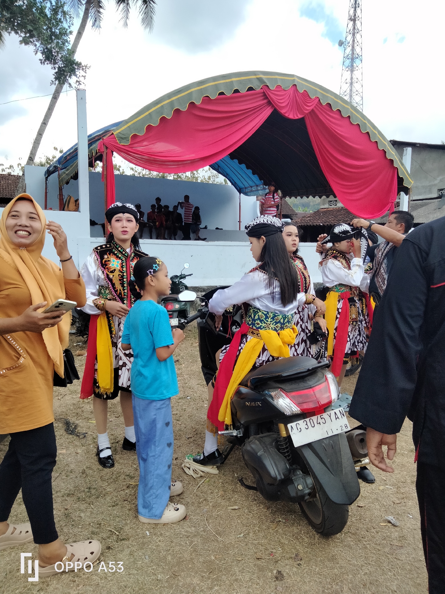 In Bendoagung, Indonesia, on a dry, grassy outdoor ground, a group of people are gathered near a stage with a red and yellow tent. A woman in a yellow headscarf and orange top holds a phone. Several young women in traditional attire with elaborate headbands and colorful sashes stand near a black motorcycle. One young woman in a blue t-shirt and blue pants stands next to the motorcycle. In the background, people are seated on bleachers under the tent, and a cell tower is visible against a cloudy sky. A black vehicle is partially visible on the right side of the frame.