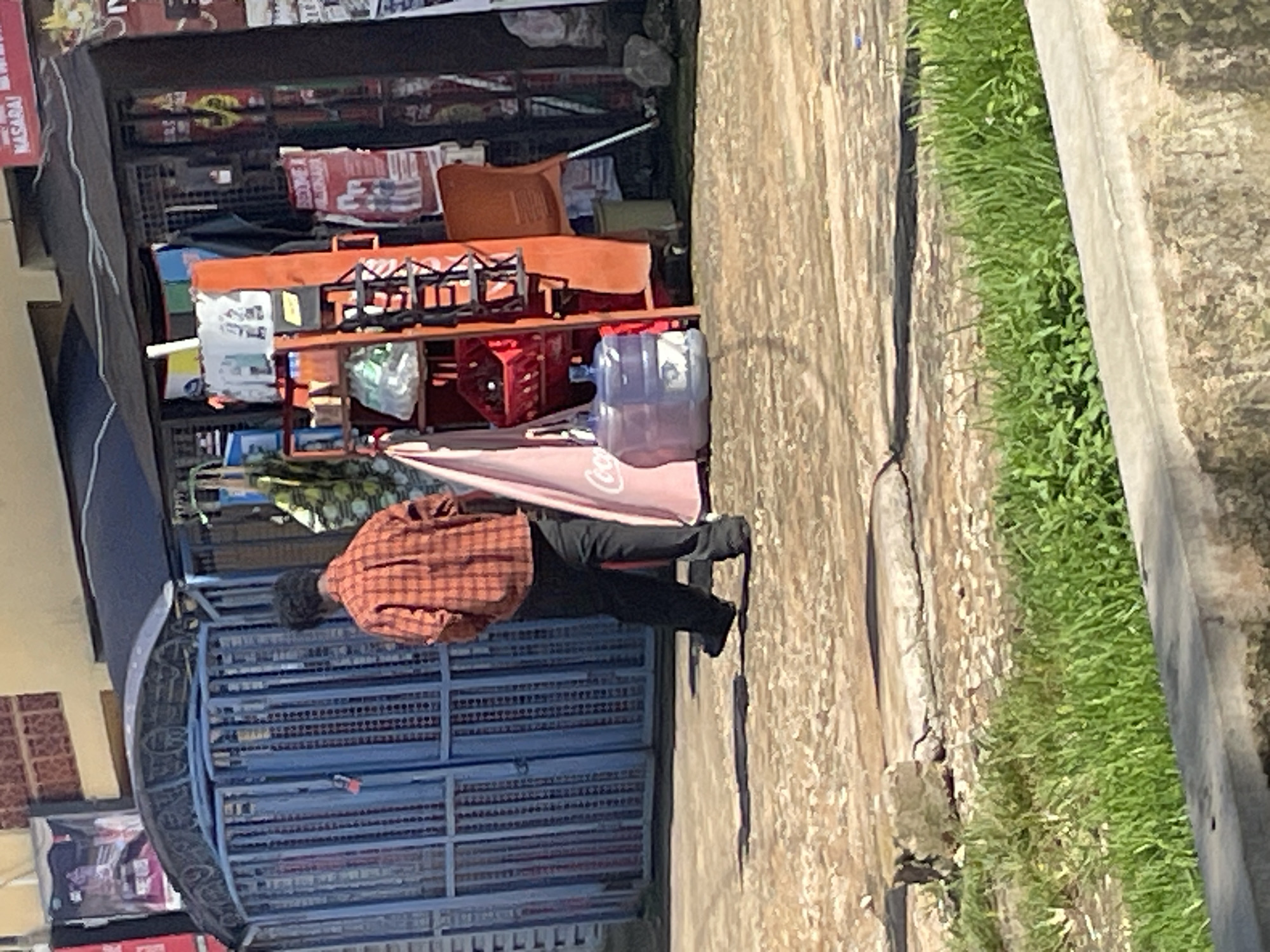The image depicts a street scene in Jos, Nigeria, with a person walking away from the camera towards a blue metal gate. The person is wearing an orange and black plaid shirt and dark pants. Behind the person, to the right, is a market stall with a black canopy. Various items are displayed on shelves and racks within the stall, including what appear to be bottles of water, baskets, and posters. An orange chair is visible near the stall. The ground in the foreground is a rough, sandy texture, with a strip of green grass bordering a concrete curb at the bottom of the frame. The overall impression is of an outdoor market area in a moderately urban setting.