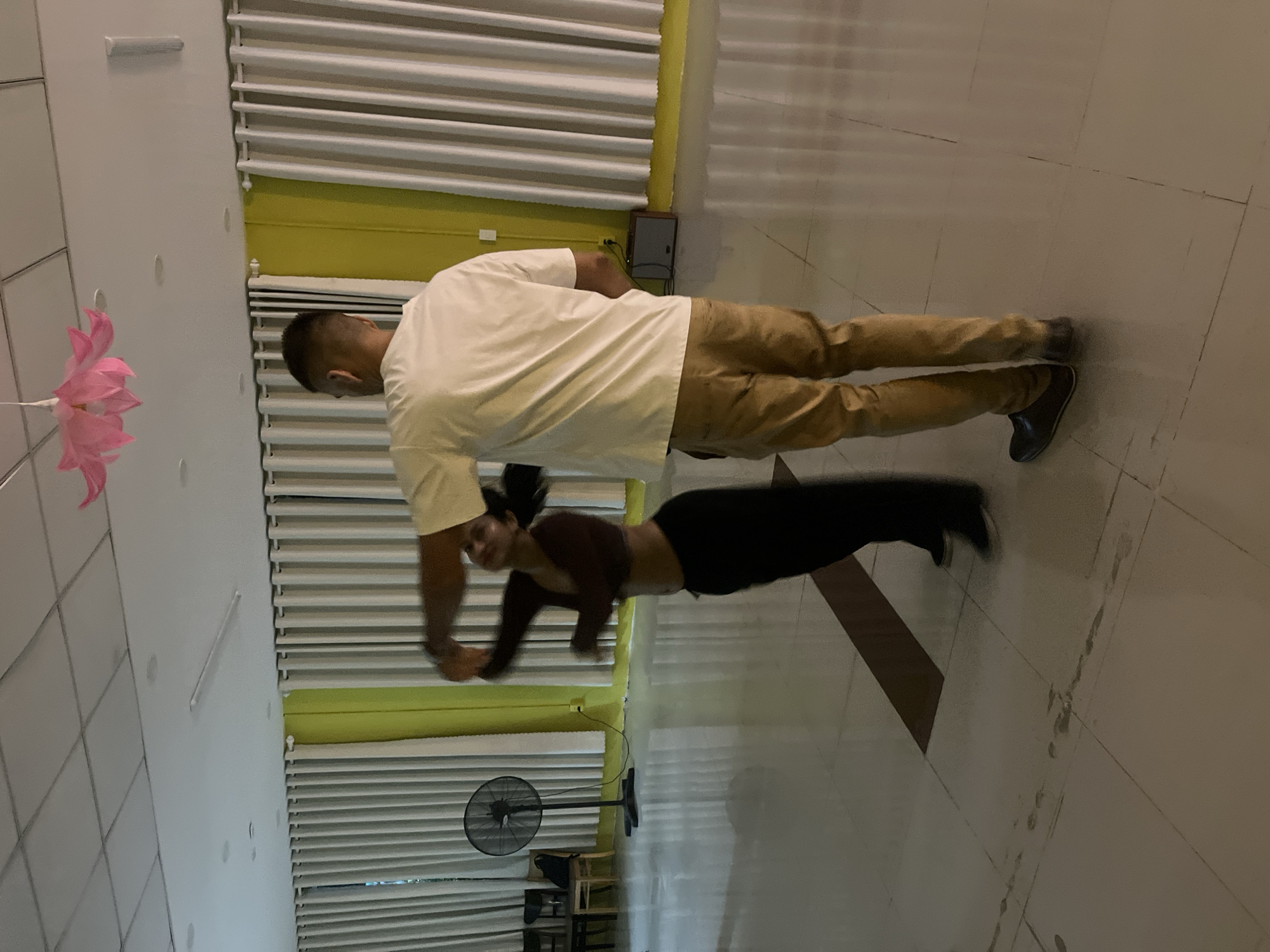 The image shows two people, a man and a woman, indoors. The man is standing on the right, wearing a light-colored t-shirt and khaki pants. He is looking down and appears to be interacting with the woman. The woman is on the left, in a plank position on the floor, wearing a dark, long-sleeved crop top and dark pants. Her body is positioned horizontally in the frame.
The setting appears to be a large room with white tiled floors and walls. There are large windows or wall panels with horizontal blinds covering them. A yellow band runs along the top of some of these panels. A pink paper decoration hangs on the wall to the left. In the background, a black oscillating fan is visible on the floor, along with some chairs and a table.
The time of day is not explicitly indicated, but the indoor lighting suggests it could be daytime or evening. The location cues, such as the fan and the general aesthetic, are consistent with a commercial or recreational space in Đà Nẵng, Vietnam. No visible text is present in the image. The overall atmosphere seems to be one of activity and possibly exercise or training.