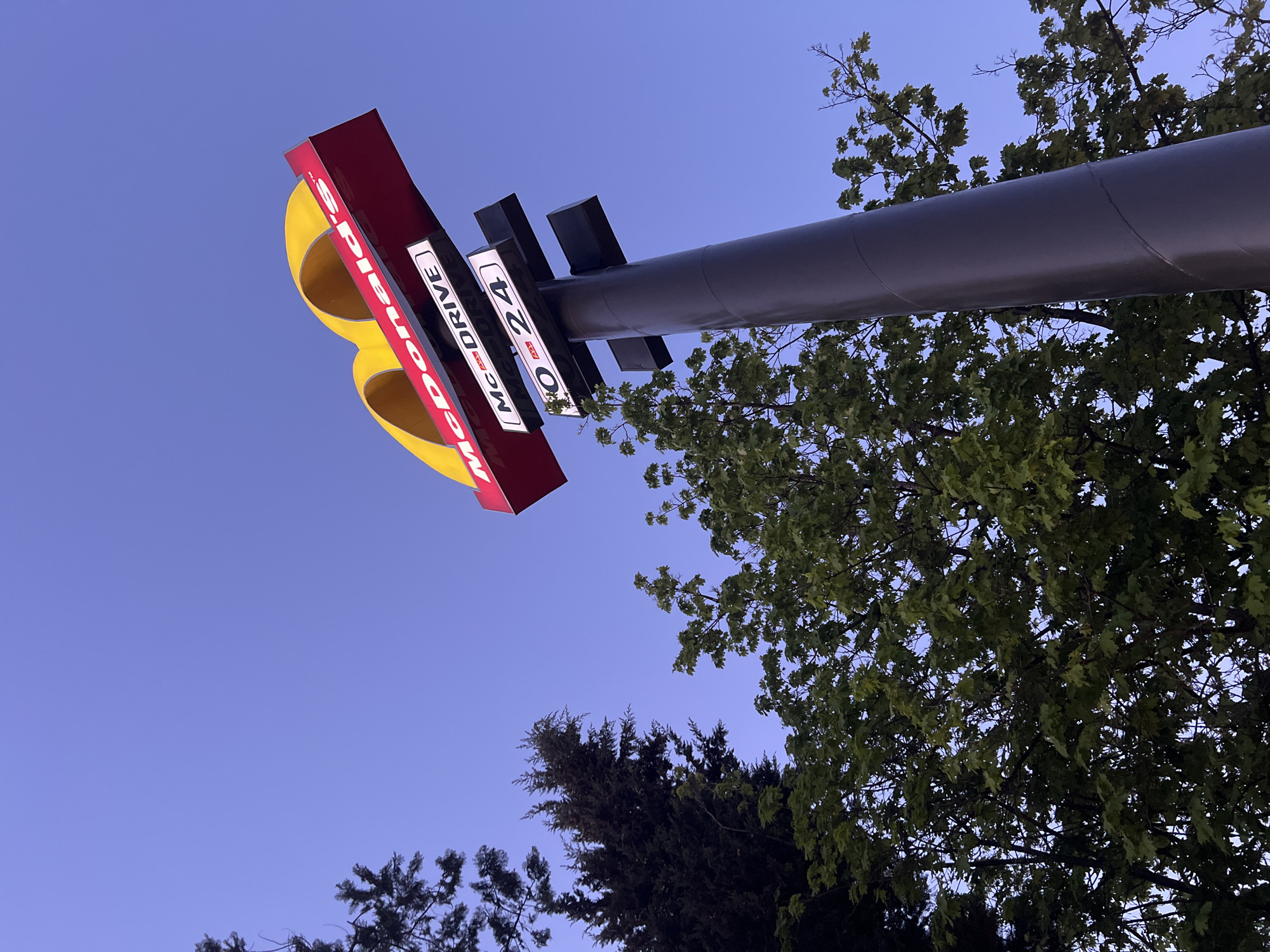The image displays a McDonald's sign captured from a low, upward-looking angle against a clear, deep blue sky, suggesting either twilight or dawn. The large sign is predominantly red, featuring the iconic golden arches in yellow, which are partially visible on the left side and appear rotated due to the camera's perspective. The sign is supported by a thick, dark grey pole that extends horizontally across the upper right portion of the frame.
Lush green foliage from several trees, possibly deciduous and coniferous, frames the right and bottom-left edges of the image, contrasting with the clear sky. The artificial illumination of the sign stands out against the natural light of the sky, indicating a time when the sun is not directly overhead.
Visible text on the sign includes "McDonald's" printed vertically on the red section, which appears rotated and partially obscured. Below it, on a rectangular white panel, "McDRIVE" is clearly legible, also oriented vertically. Adjacent to "McDRIVE", another white panel shows "NO 24" in black lettering, accompanied by a small red graphic. This likely signifies "open 24 hours" for the drive-through service. No human figures or activities are present in the static scene.