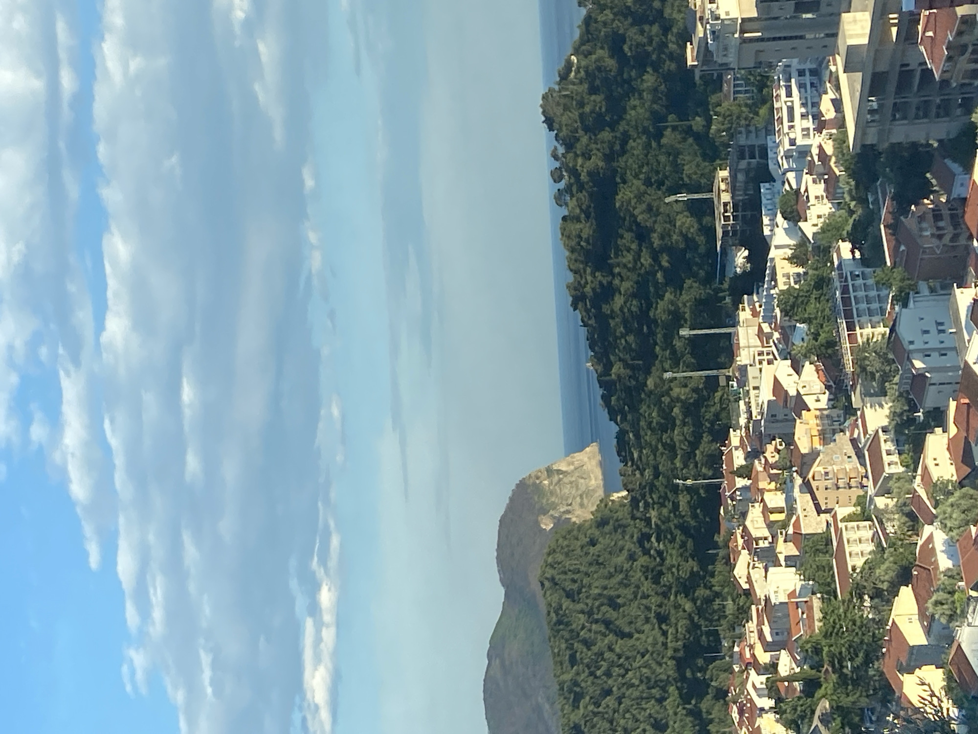 The image shows an aerial view of a coastal town in Montenegro, likely near Nerin. The scene includes a blue sky with scattered clouds, a narrow stretch of ocean, and a rocky hill covered in green vegetation. On the right side of the image, there is a dense cluster of buildings with red-tiled roofs, interspersed with trees. There is a construction site in the town. The image appears to be taken from a high vantage point, possibly from a tall building or a drone.