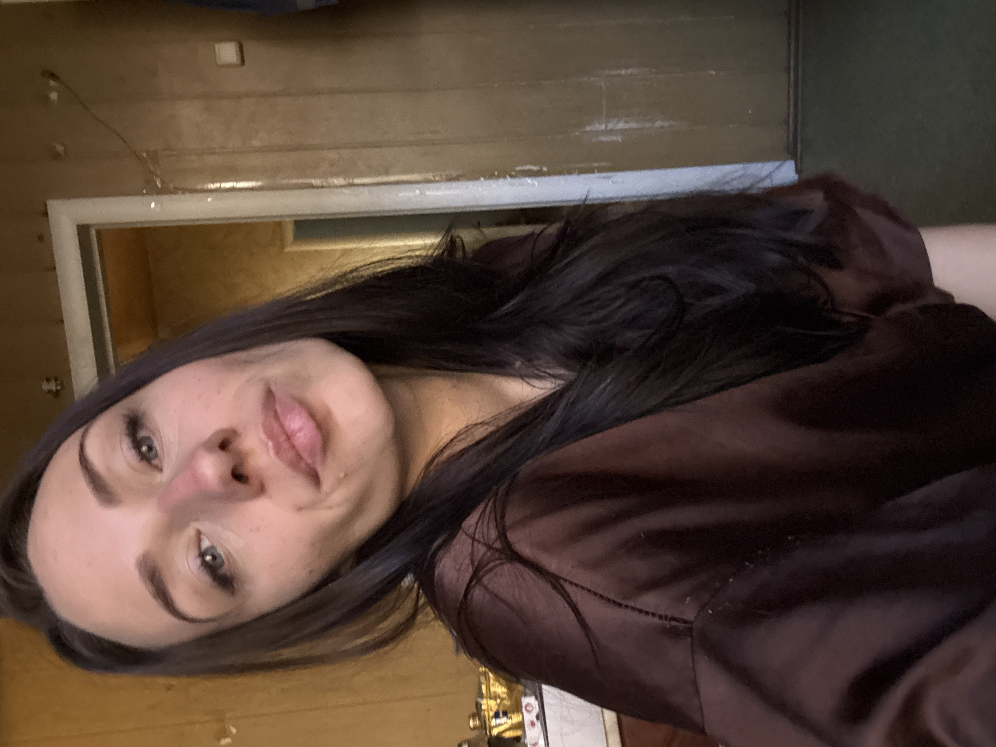 A woman with fair skin, dark hair, and green eyes is centered in the image, looking slightly towards the left. She is wearing a brown, silk-like top. The background consists of a room with light-colored wooden walls, a doorway, and a glimpse of what appears to be a counter with items on it. The location is likely indoors and given the context of the prompt, perhaps the interior of a building. Without additional information, it is not possible to determine whether it is in the city of Saint Petersburg, Russia.