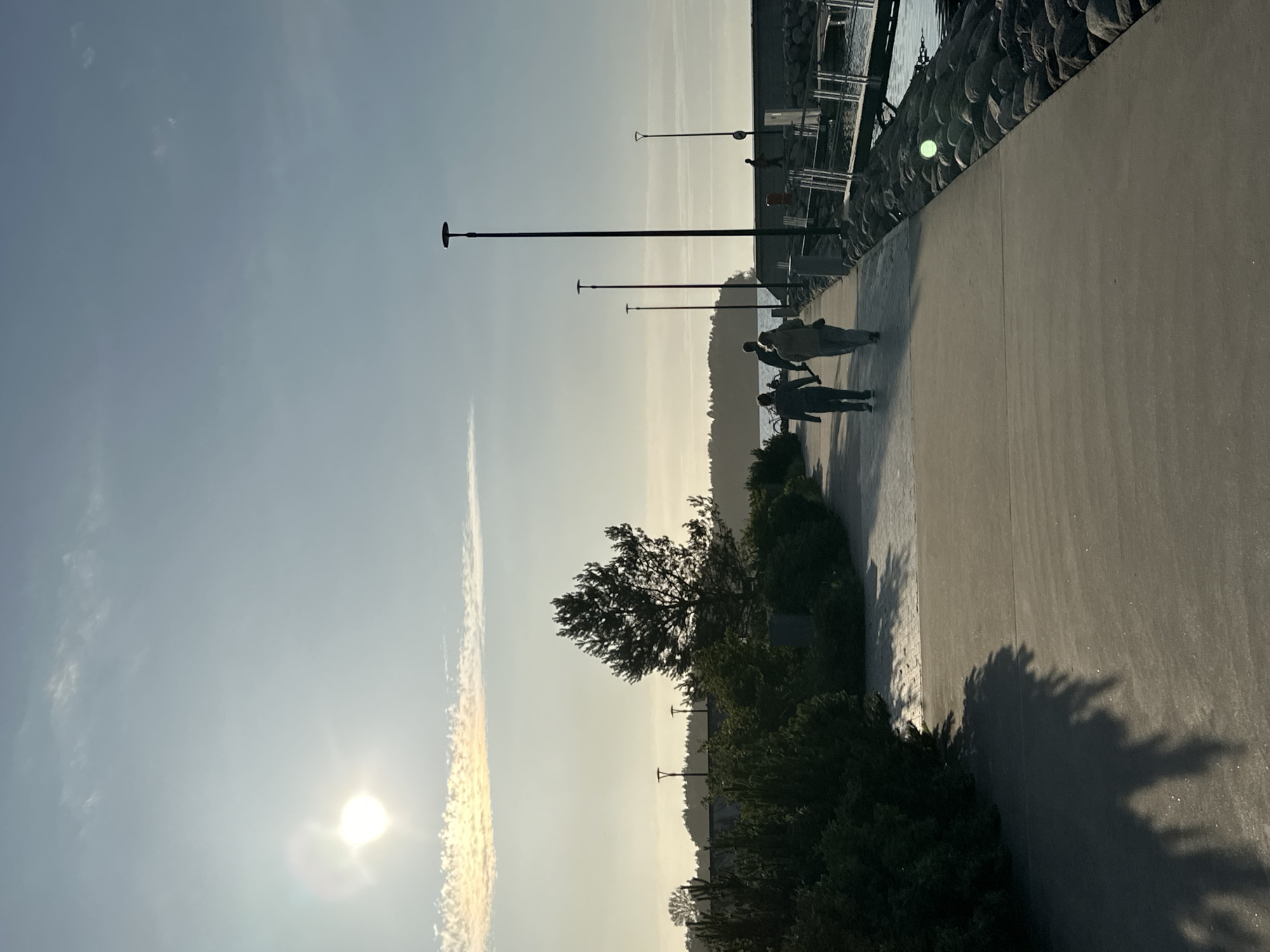 The image depicts a paved walkway bordering a body of water. The walkway is lined with bushes on the left and a stone embankment on the right, leading to a pier. Several lampposts are visible along the walkway. In the distance, a tree stands against a hazy sky with a bright sun. The sky is clear with some wispy clouds. The overall lighting suggests it is either early morning or late afternoon. Three individuals are walking away from the camera on the walkway. The environment appears to be a scenic waterfront area, possibly a promenade or park. The location is not explicitly identifiable as Tallinn, Estonia.