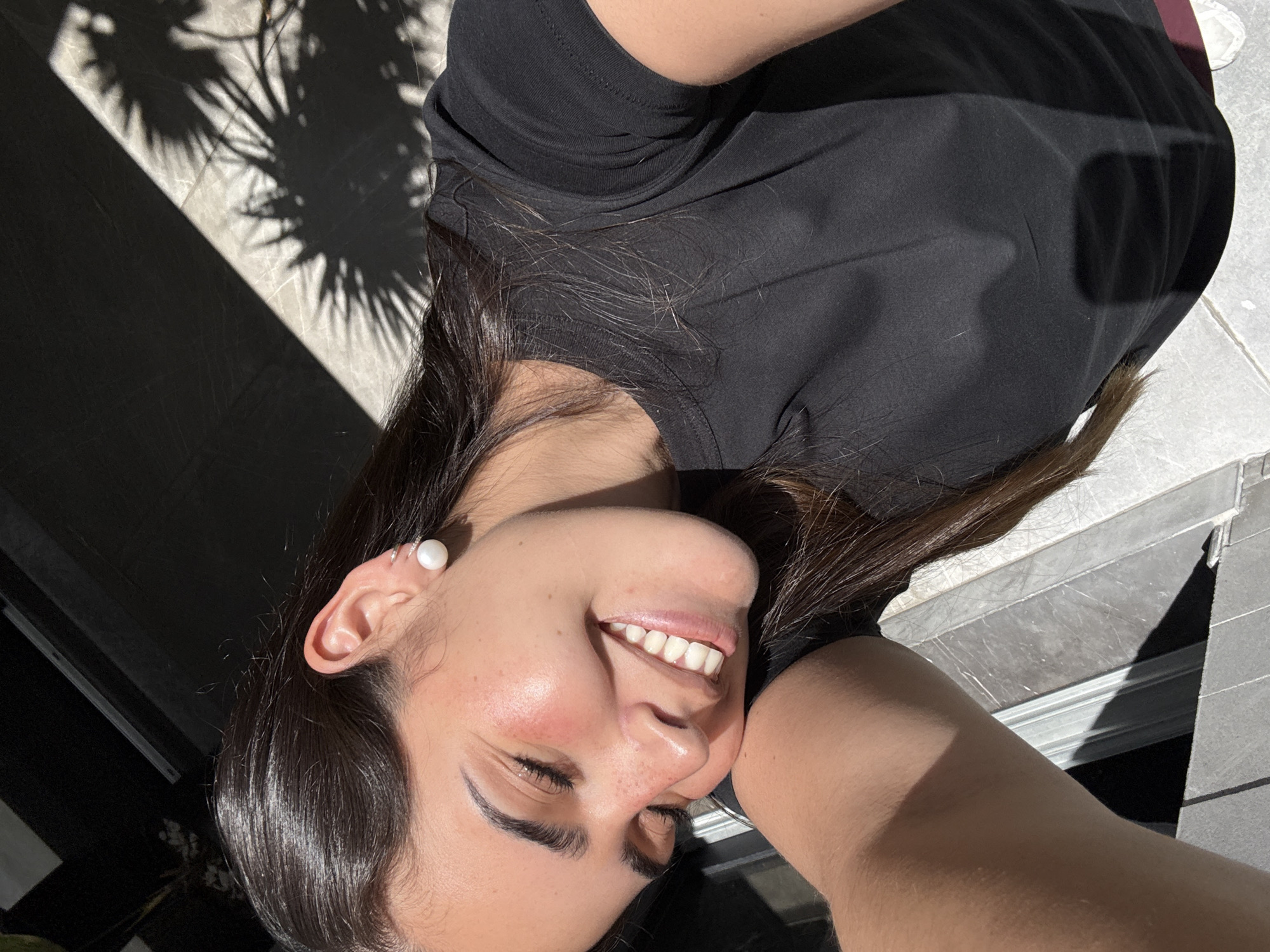 A woman is visible in this close-up, angled view. She is smiling, and her skin appears to be sun-kissed. She is wearing a black T-shirt and an earring. Part of the ground is visible, and there are shadows cast on it. The location is San Bernardino Tlaxcalancingo, Mexico.