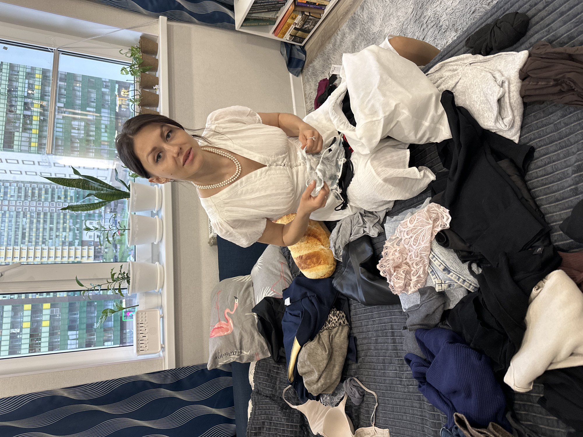 A woman with dark hair and fair skin, wearing a white dress and a pearl necklace, sits amongst a pile of clothes on a bed with blue and grey striped bedding. She is holding a black and white garment. Among the clothes are items of various colors including white, grey, black, blue, and pink. A pillow with a pink flamingo design is visible. A plush bread pillow lies beside her. In the background, a window reveals a view of multi-story buildings, indicative of an urban setting. Plants in white pots are on the windowsill. A bookshelf filled with books is also visible in the background. Based on the buildings visible, the location is likely Murino, Russia.