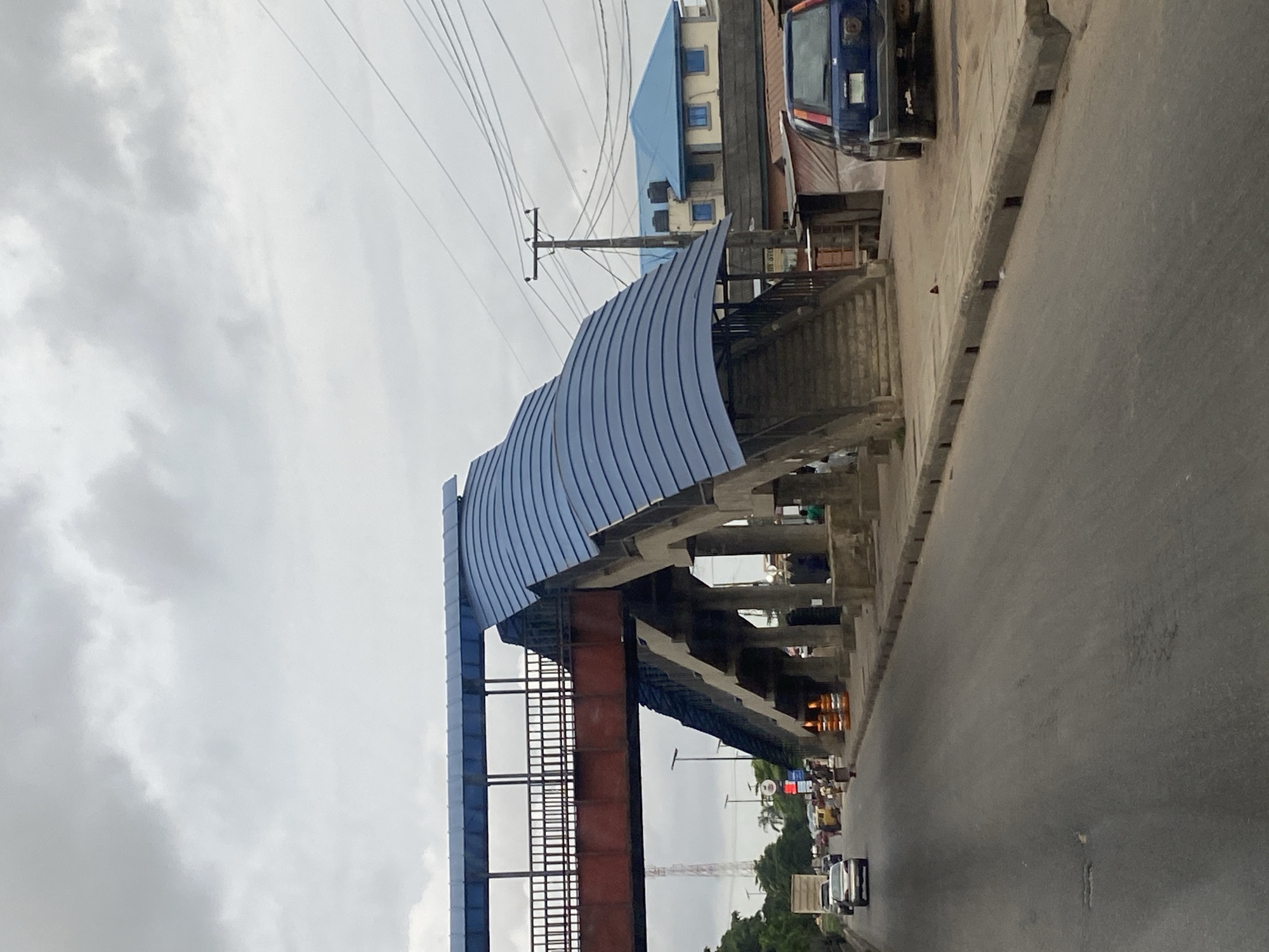 An outdoor, eye-level, medium shot captures a pedestrian bridge over a busy road in Lagos, Nigeria. The sky is overcast with gray clouds. The pedestrian bridge features a red metal walkway with a blue, curved, metal roof and concrete supports. The stairs of the bridge are visible, leading down to the sidewalk next to the road. Several vehicles, including a blue SUV, a white sedan, and a truck, are on the road. A few pedestrians are seen on the sidewalk and near the bridge supports. Buildings and utility poles are visible in the background. The road is paved, with a concrete curb and a drainage system along the side. The overall scene depicts a typical urban environment in Lagos, characterized by its bustling traffic and infrastructure.