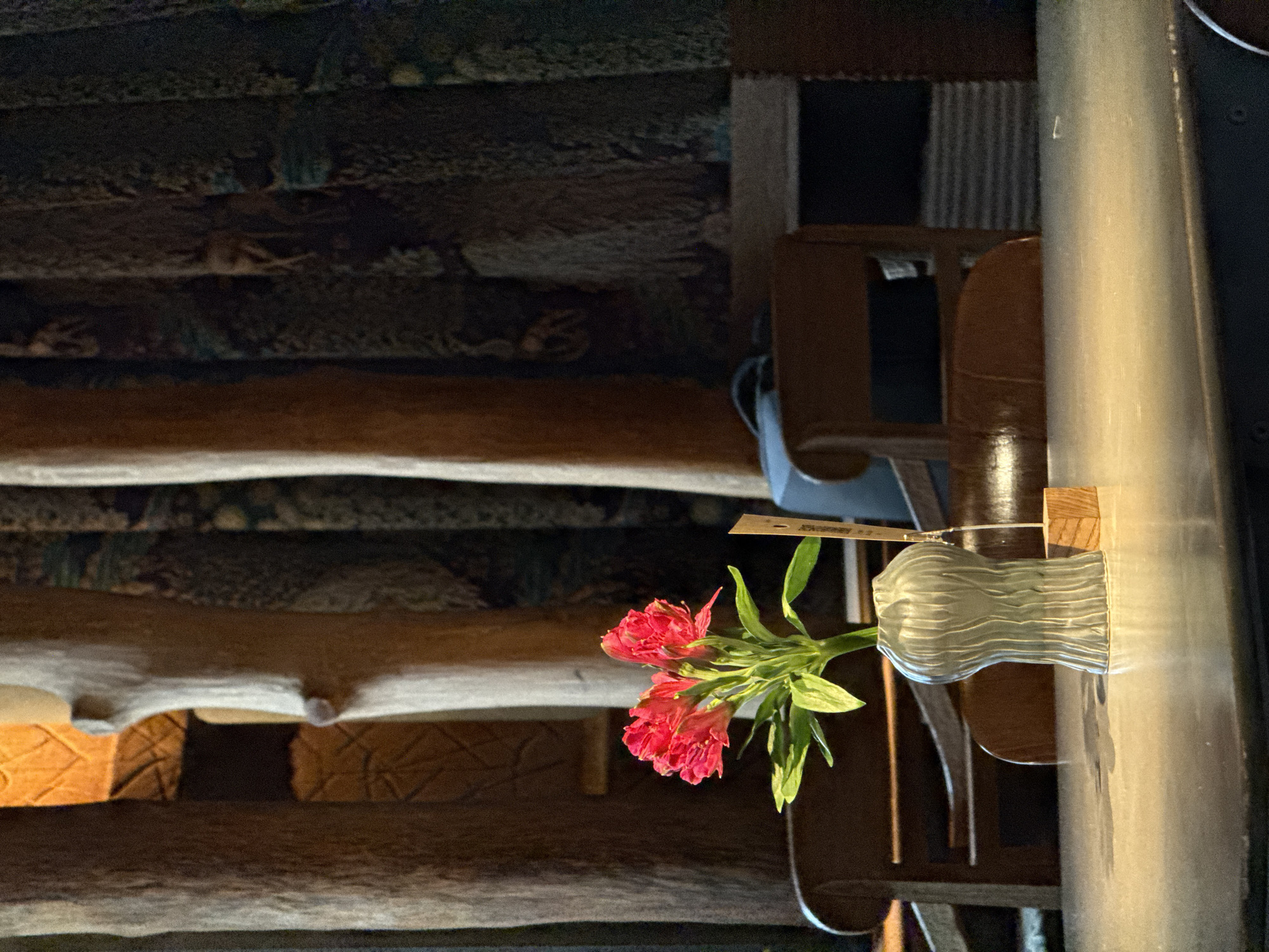 The image shows a table with red flowers in a vase, set against the background of a textured wall. The vase sits on a wooden table next to a metal pole. A wooden chair is also visible. While the image is evocative, there's insufficient data to definitively determine if it's in Kazan, Russia, or any other specific location.