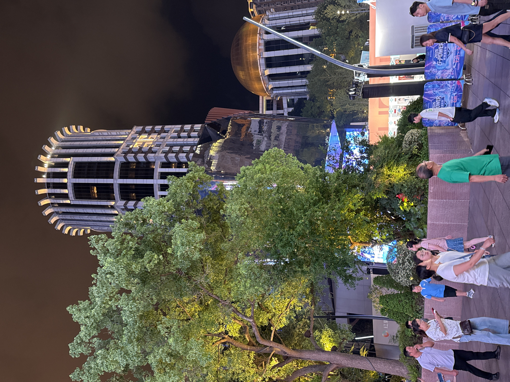 The image depicts an outdoor evening scene in Shanghai, China. A distinctive cylindrical skyscraper with illuminated top accents is prominently featured against a dark, cloudy sky. To its right, a building with a golden dome and fluted columns is visible. In the foreground, a large, leafy tree is illuminated from below. Several individuals are walking along a paved area, some are engaged in conversation, and others are looking at their phones. The ground is paved with rectangular stones. In the background, there are more buildings, trees, and some illuminated signage. A low wall with a textured surface runs horizontally across the midground. The overall atmosphere suggests a bustling urban environment at night.