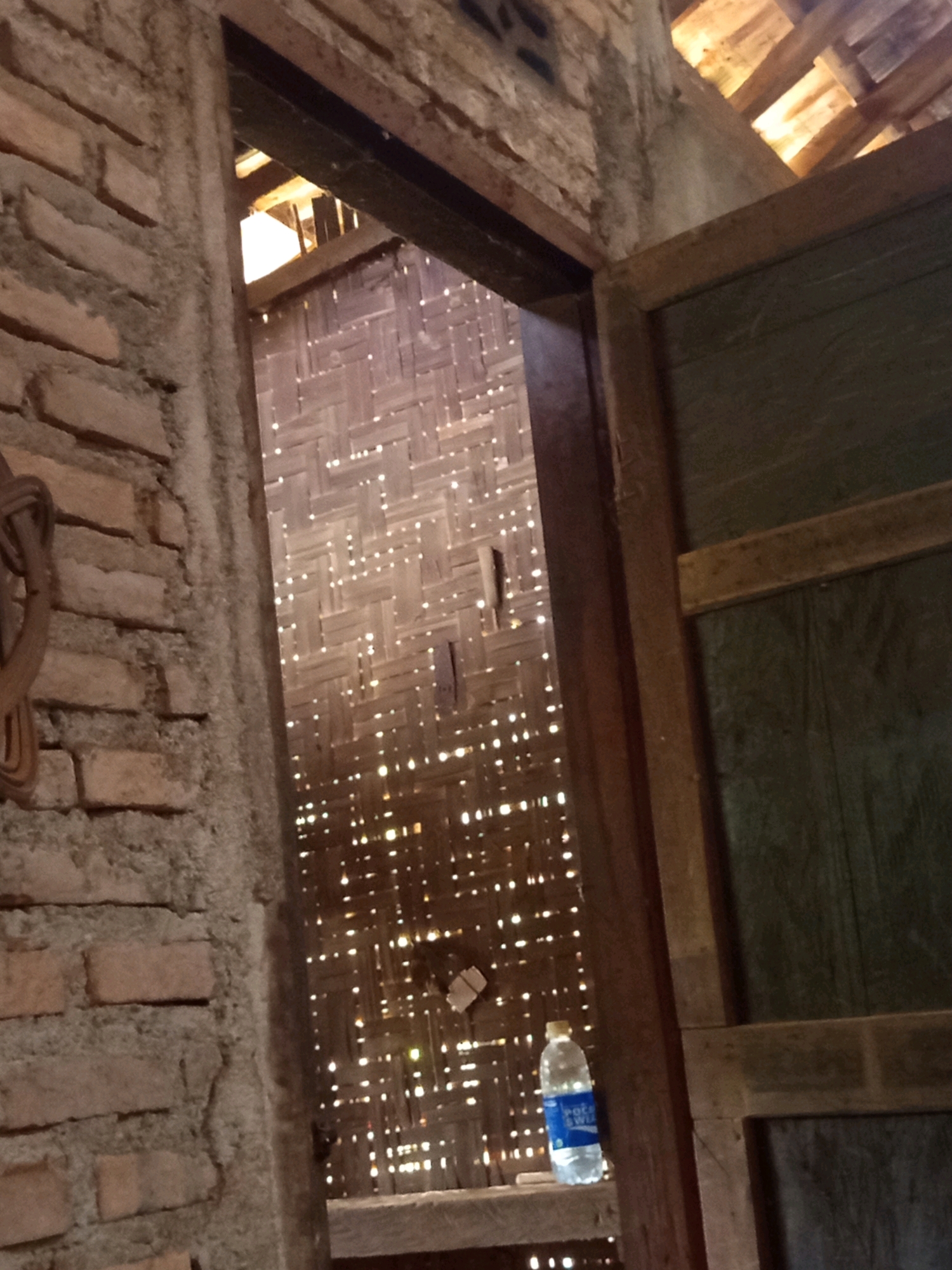 The image shows a view through a doorway into a room with a woven bamboo wall, likely in Karang Sari, Indonesia. The doorway is framed by rough brickwork on the left and a wooden door frame on the right. The woven bamboo wall behind the doorway is illuminated by light filtering through its many small openings, creating a speckled pattern. A plastic water bottle with a blue label is visible on a shelf in front of the bamboo wall, near the right side of the doorway. The ceiling above the doorway is constructed with wooden beams and planks, with light emanating from above. The overall impression is of a simple, possibly rustic structure.
