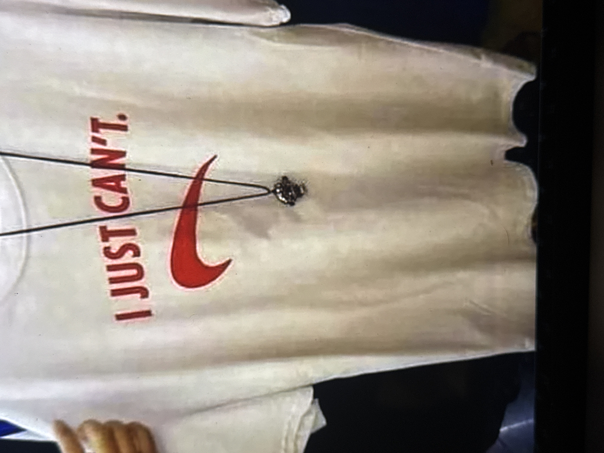The image shows a white t-shirt with red text and a red Nike logo. The text on the t-shirt reads "I JUST CAN'T". A thin black cord with a small, dark pendant is draped across the t-shirt, positioned over the Nike logo. The t-shirt appears to be made of a cotton material and is slightly creased, suggesting it might have been worn or is folded. The lighting in the image is somewhat dim and uneven, casting shadows and giving the t-shirt a slightly yellowish hue. In the bottom left corner, a person's hand is partially visible, with fingers extended. The background is dark and out of focus, making it difficult to discern any specific setting or location cues. The overall presentation of the t-shirt and the pendant suggests it might be a piece of apparel with a personal or symbolic meaning. The provided location context of Kuta Selatan, Indonesia, does not directly inform the visual elements of the image itself.