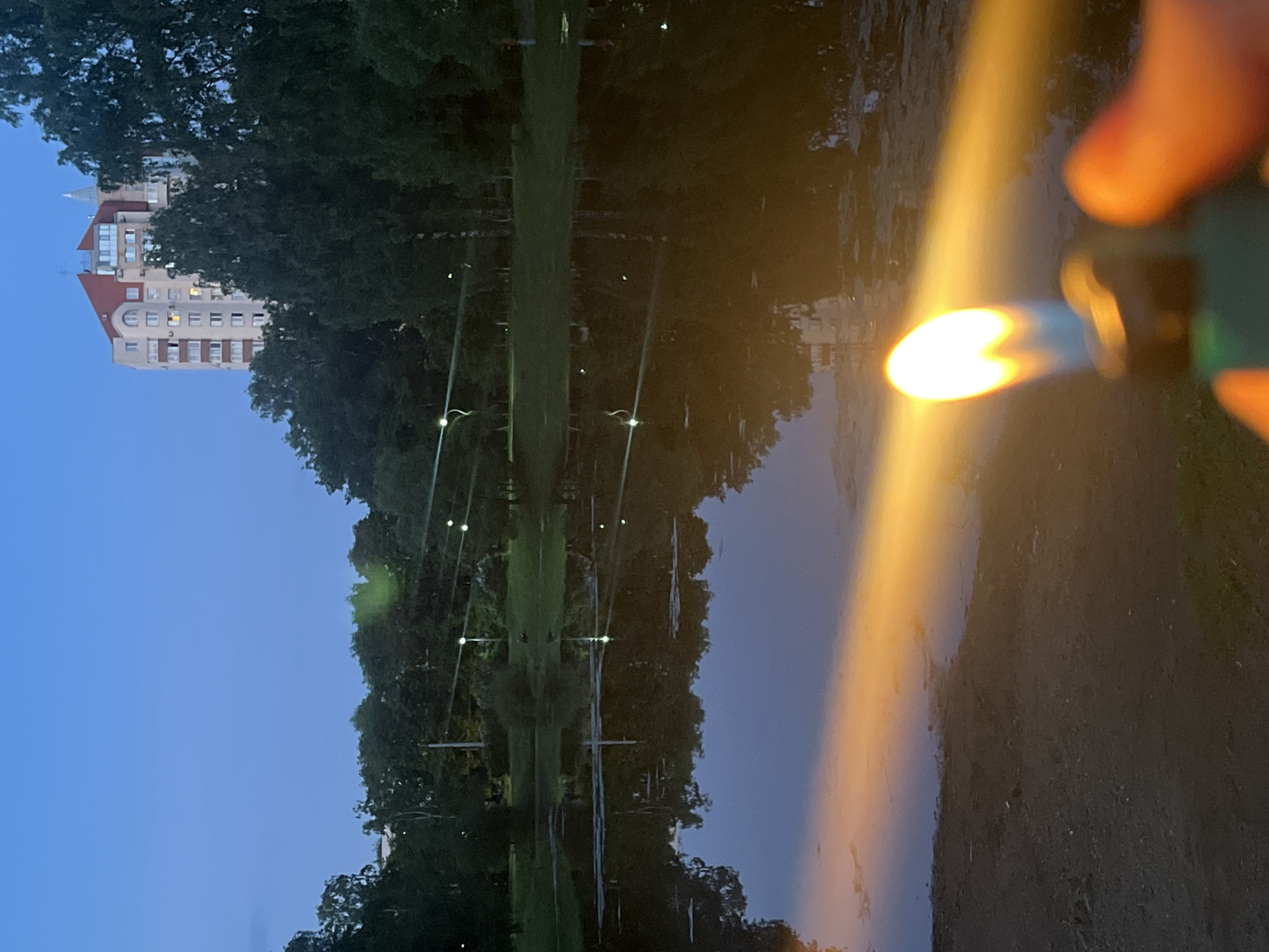 This image captures an outdoor scene at twilight or early night in the city of Sinyavino, Russia. In the immediate foreground, a human hand is visible, holding a green utility lighter with an active, bright orange-yellow flame. This flame projects a strong, elongated light beam across a dark, damp ground surface and onto the still water of a pond or large puddle. The water reflects the dark blue twilight sky, surrounding trees, and several distant, faint point-light sources. These background lights, appearing as small white or green dots, are also visible within the tree line and reflected on the water's surface. In the background, a dense canopy of dark green trees frames a multi-story building. This structure features light-colored walls, a prominent red-tiled roof with an angular design and dormer windows, and several illuminated interior windows. A small spire-like fixture is visible on the building's roof. The sky above is a clear, deep blue, indicative of the time of day.