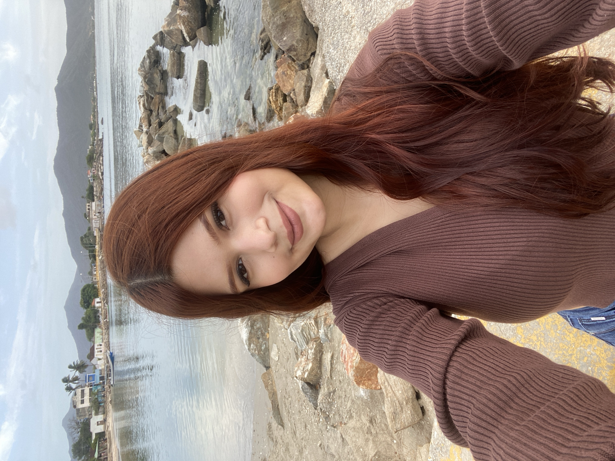 A young woman with long, auburn hair takes a selfie on a rocky shoreline. She is wearing a brown ribbed sweater and blue jeans. The background shows a calm body of water, with a small town and hills in the distance. The sky is overcast. The scene is likely Juan Griego, Venezuela, a coastal town known for its scenic beauty.